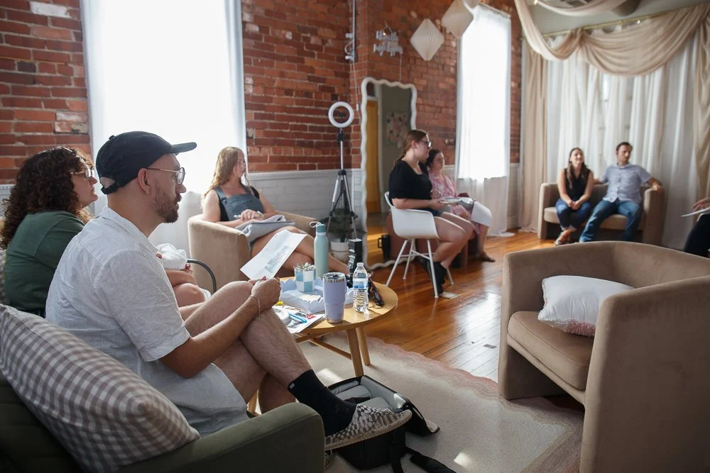 Modern wooden house overlooking a lake with mountains in the background, surrounded by grass and trees, under a cloudy sky.