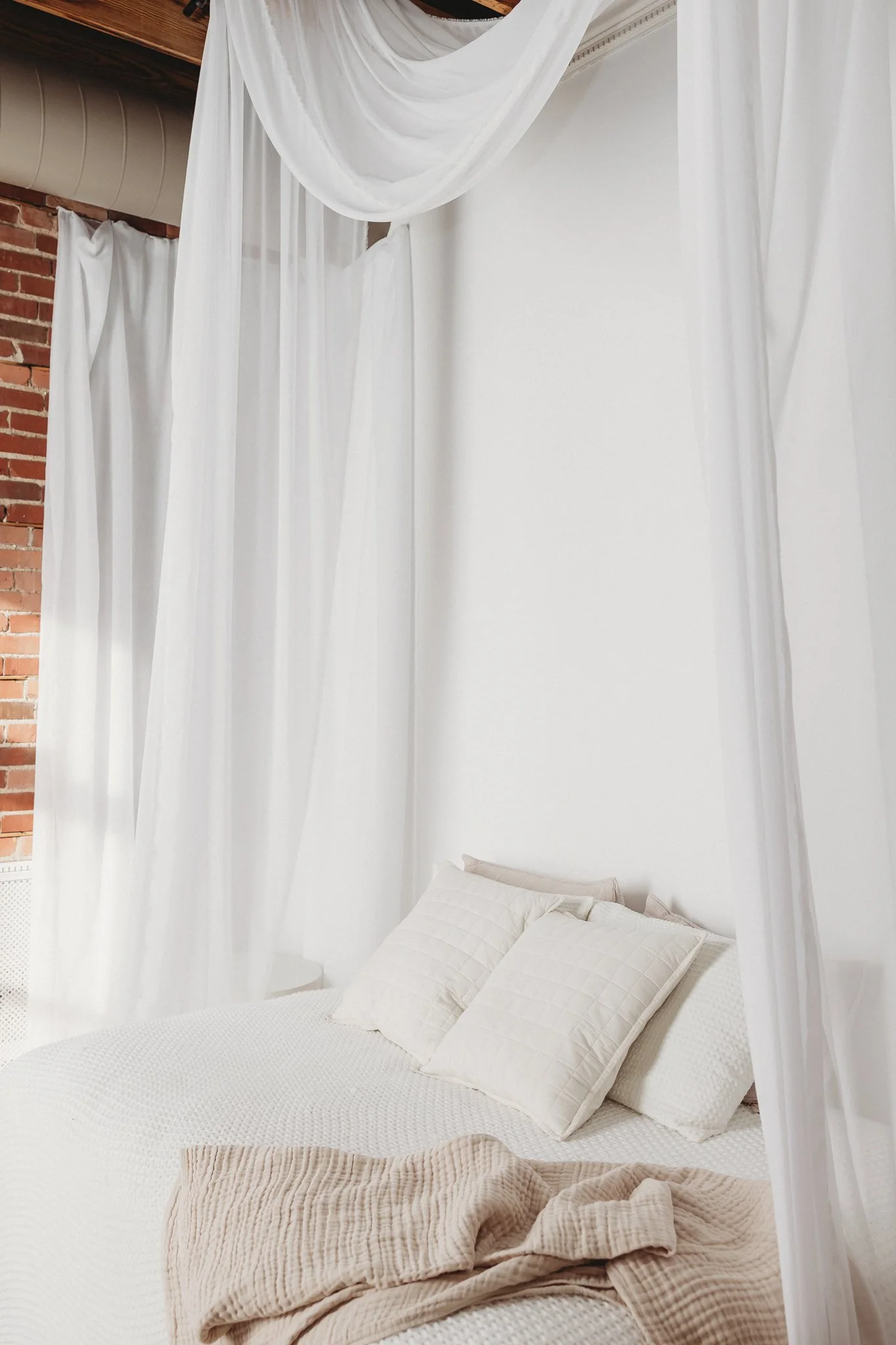 A bed with white pillows and a beige quilted blanket under white sheer curtains in a bedroom with exposed brick walls.