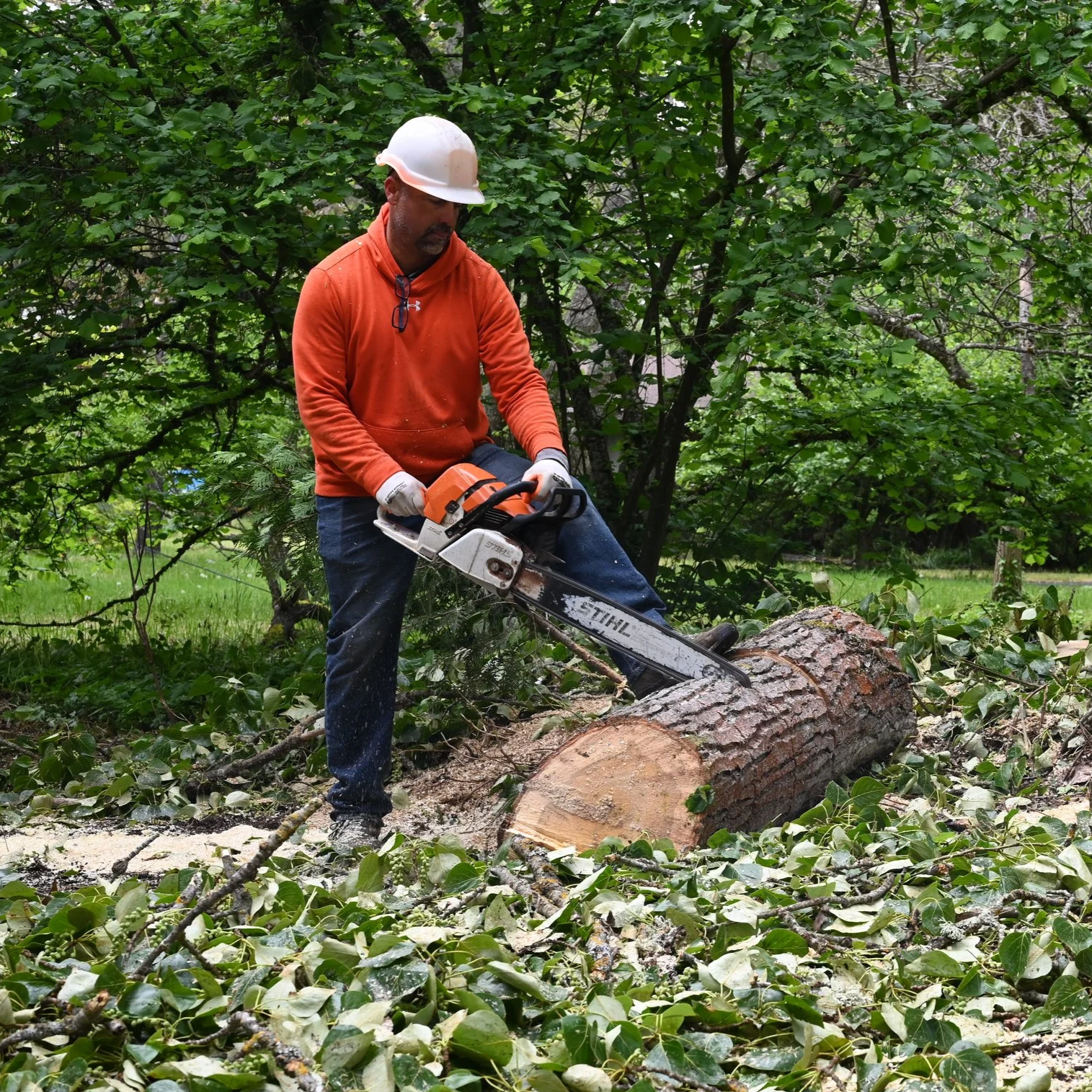 Man wearing an orange hoodie and a yellow hard hat standing outdoors with trees in the background.