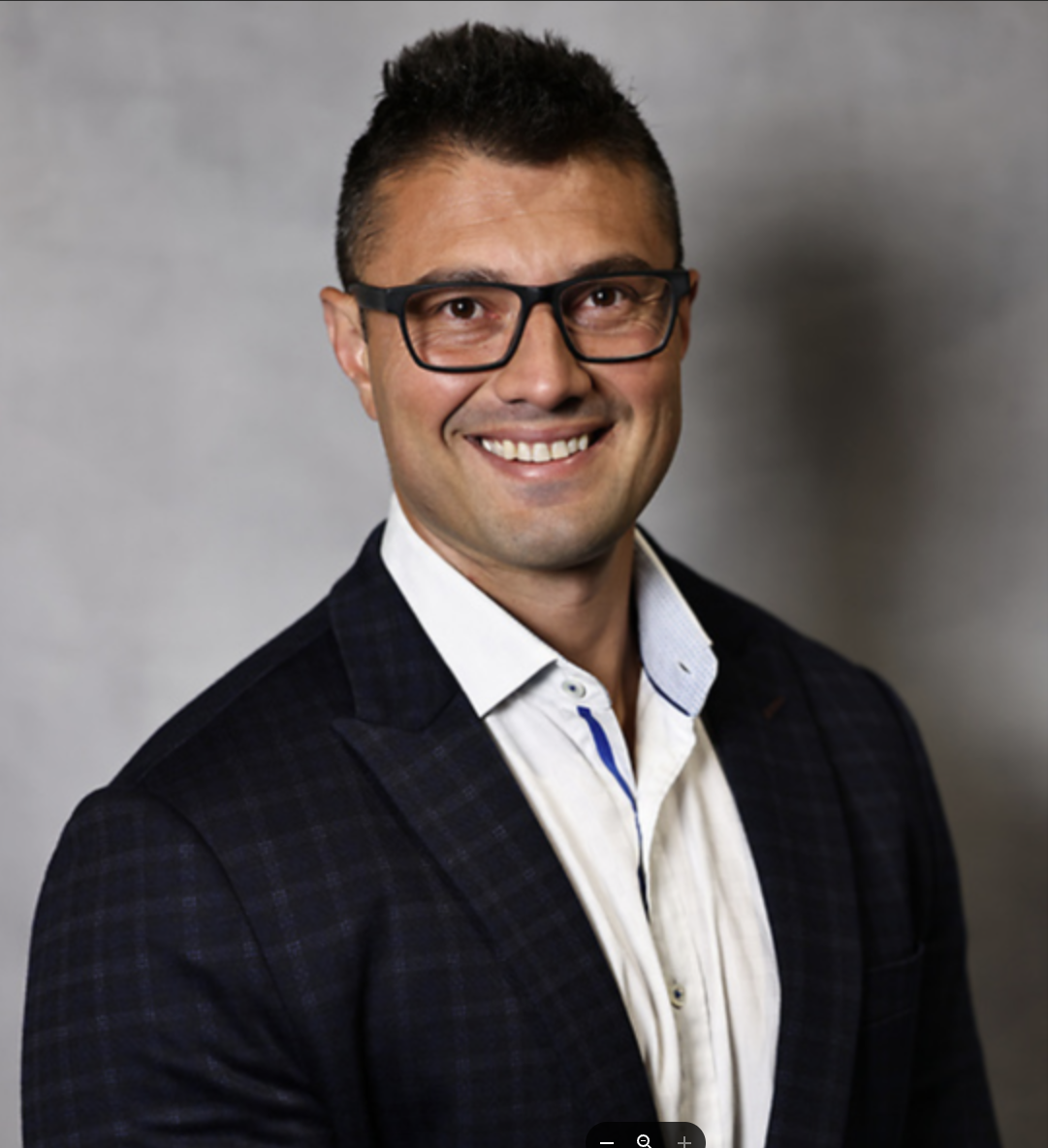 A smiling man wearing glasses, a white shirt, and a dark blazer against a gray background.