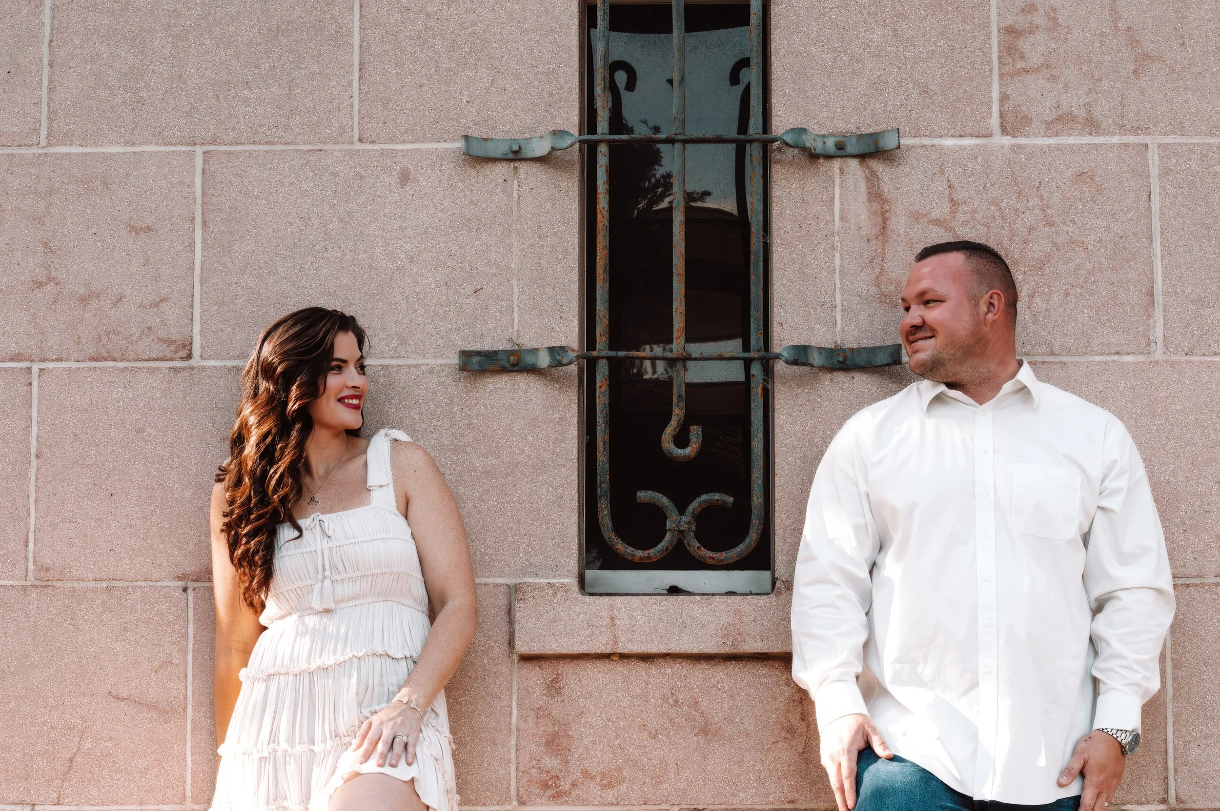 A couple smiling at one another while standing against a stone wall with a narrow window between them, photographed in natural light.