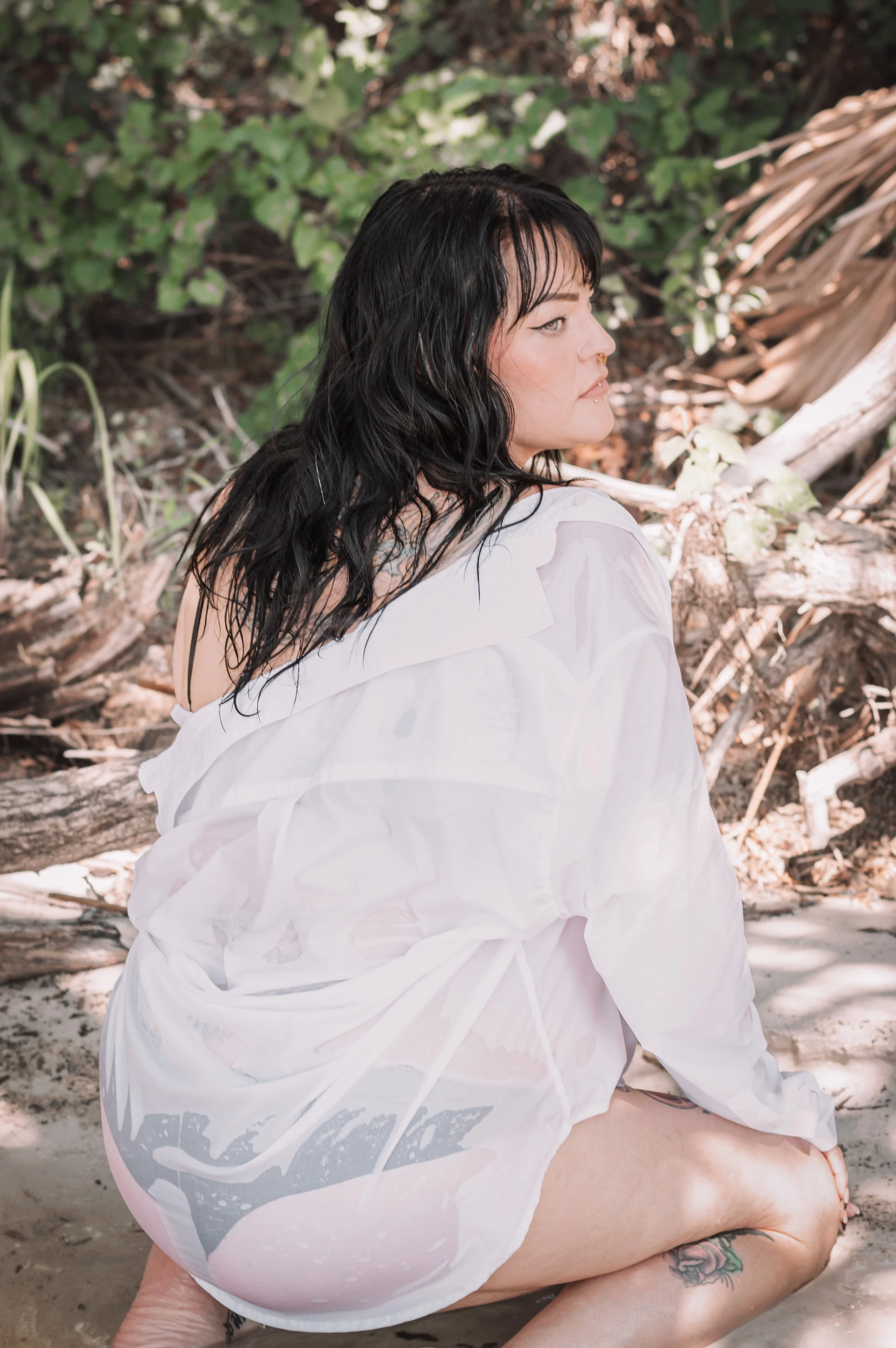 A woman seated on a sandy beach in a sheer white shirt, surrounded by greenery and driftwood in a natural coastal setting.