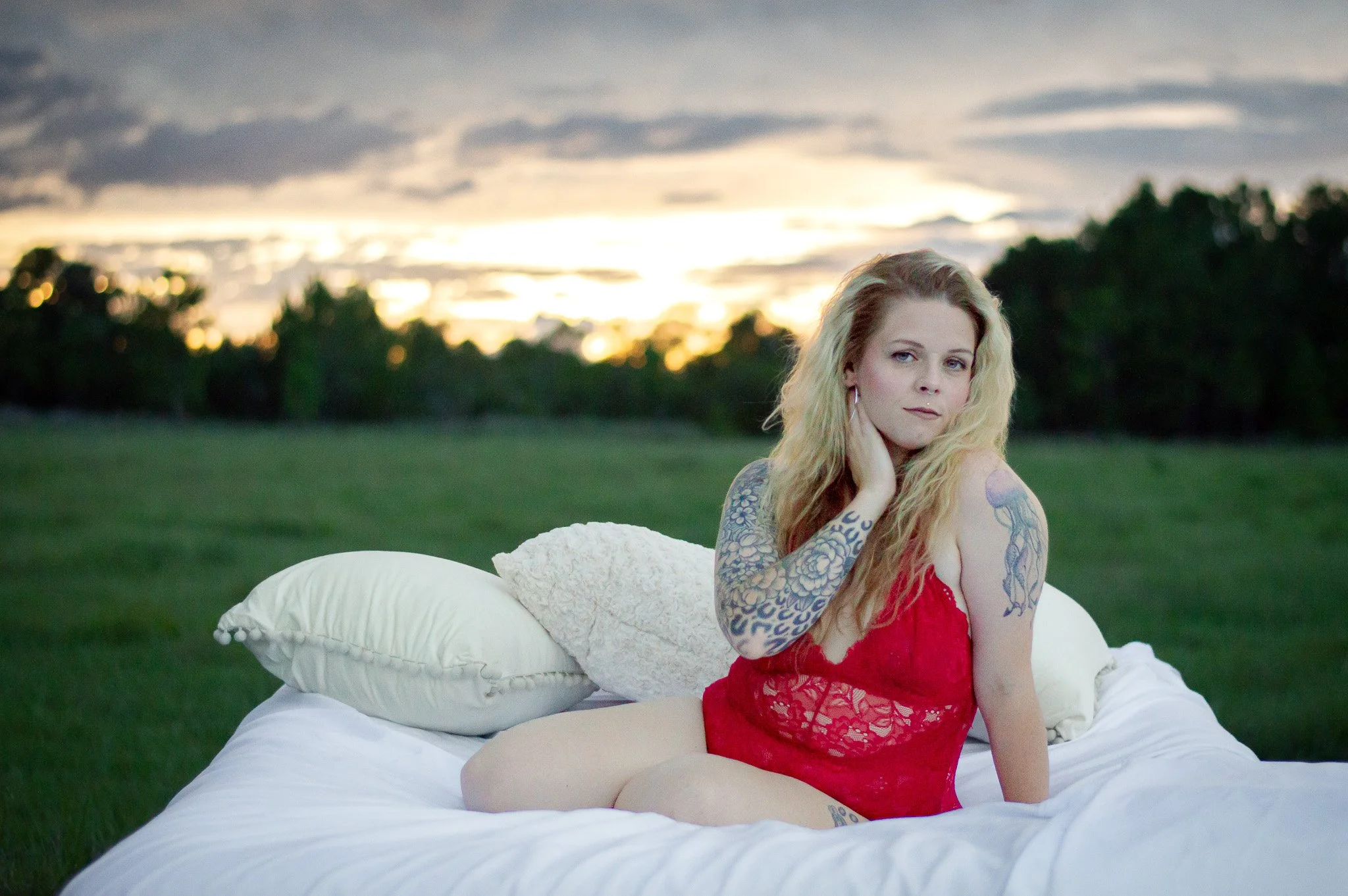 A woman wearing a red lace lingerie seated on a white bed outdoors in a field at sunset, photographed in soft natural light.