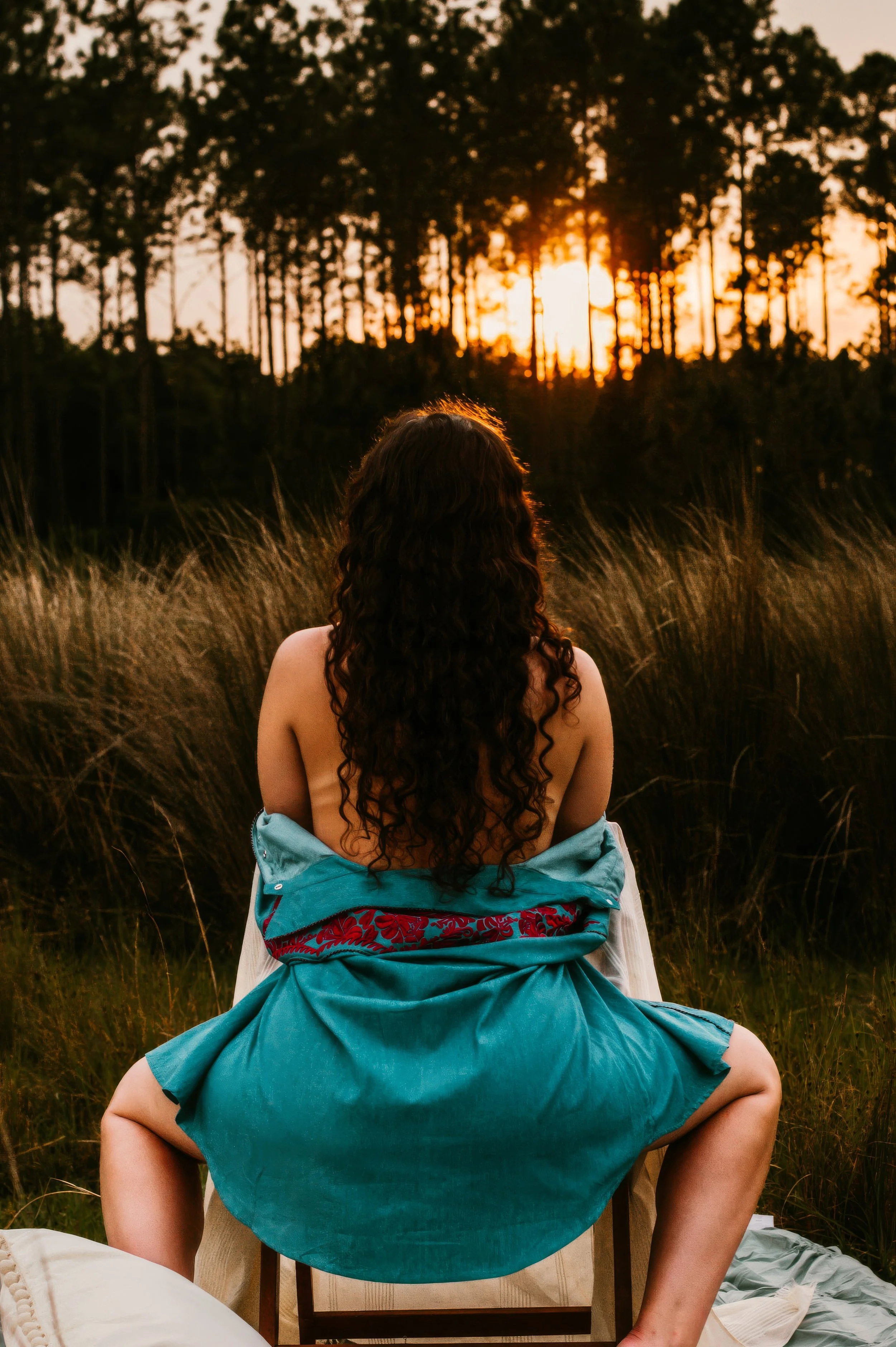 Boudoir portrait of a woman seated outdoors at sunset, wrapped in fabric, surrounded by tall grasses and a wooded landscape.