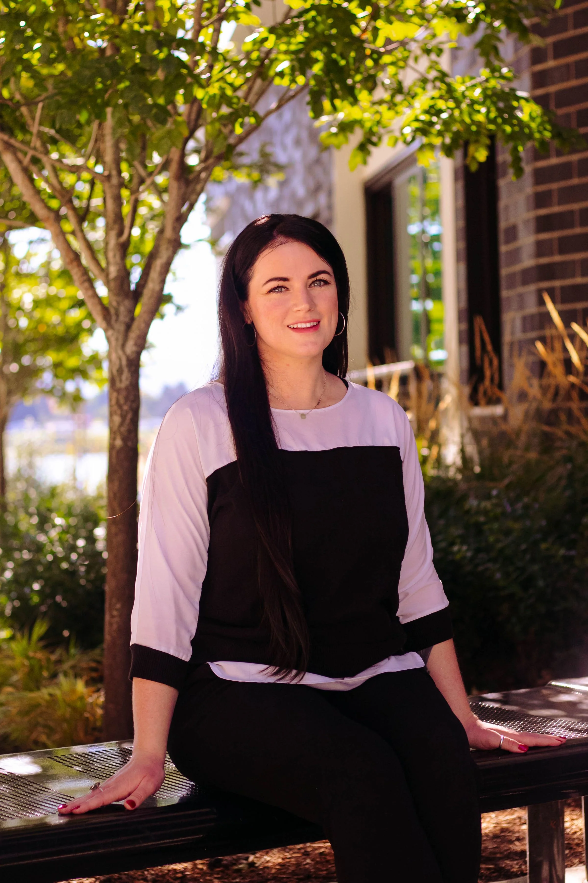 A practitioner with long dark hair, wearing a black and white top, sitting on a park bench under a tree, smiling. Kirkland, Washington. Vancouver, Washington. Mental health. Women's health. Fertility. Menopause. Microneedling. Cosmetic acupuncture.