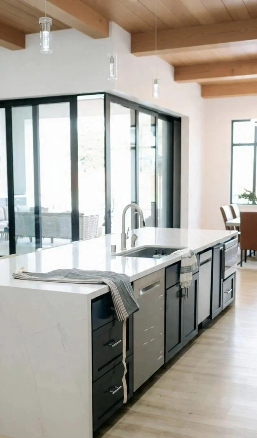 Modern kitchen with white island counter, black cabinets, stainless steel dishwasher, and hanging pendant lights, with large windows and wooden ceiling beams.