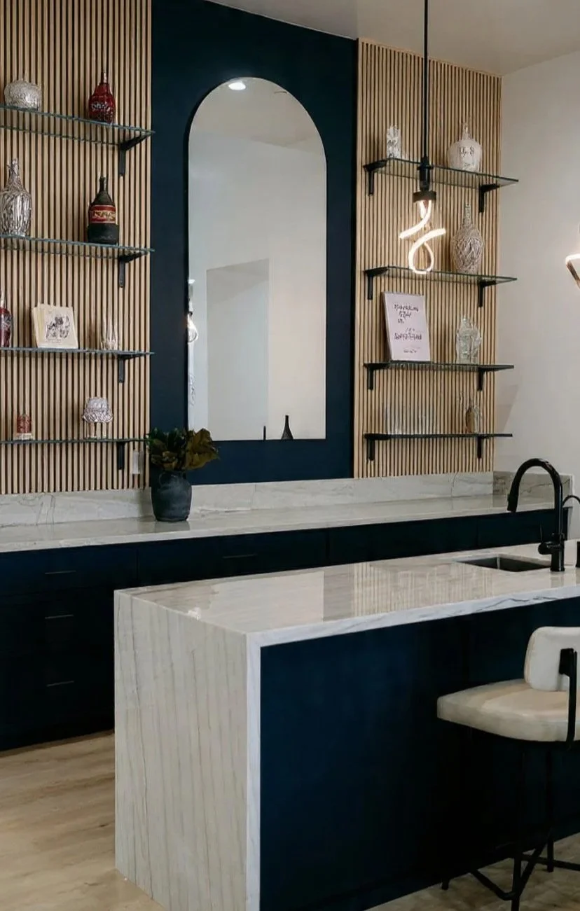 Modern kitchen with navy and wooden accents, open shelving with decorative vases and a neon light, marble countertop and cabinet, potted plant, and a stool.