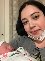Woman holding a newborn baby wrapped in a hospital blanket, smiling at the camera, with a facial mask around her neck.