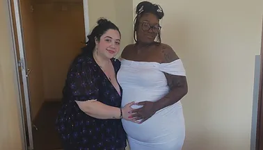 Two women standing together indoors, with one woman in a white dress appearing pregnant.