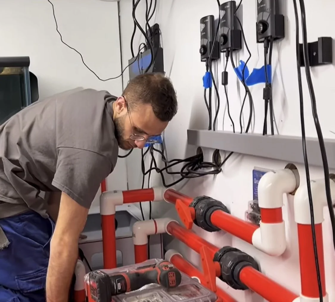 A man working on plumbing installation with red and white pipes and a drill in a utility or workspace.