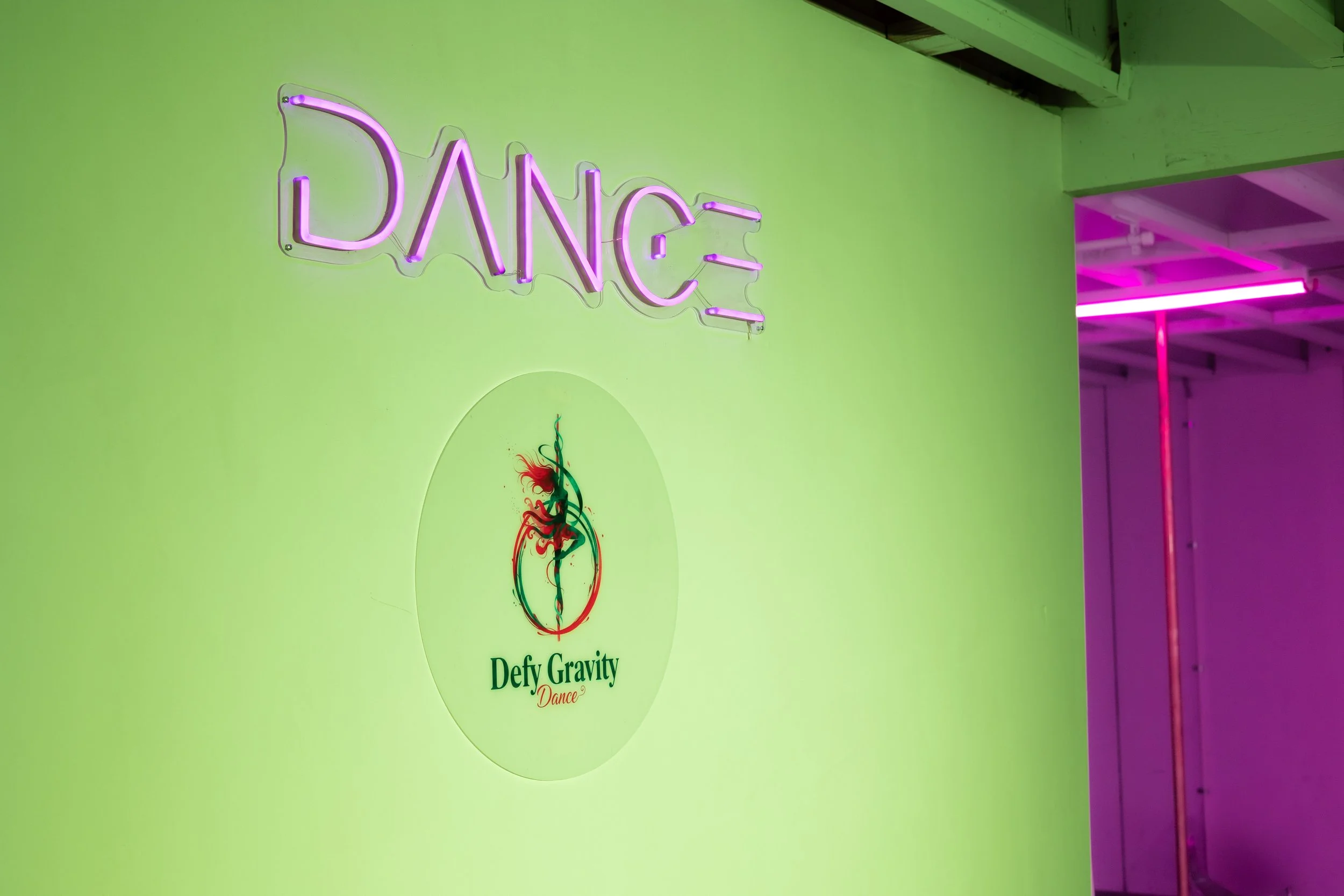 Neon sign spelling 'DANCE' in purple on a green wall, with a logo below that says 'Defy Gravity Dance' and an abstract figure of a dancer in red and green.