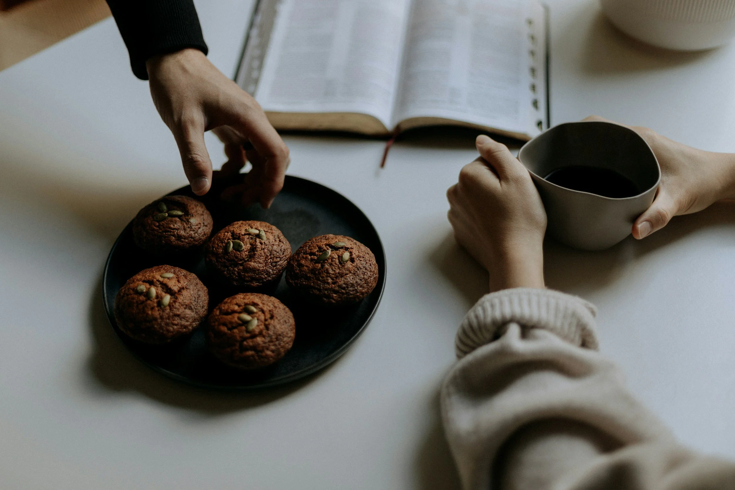 A person is reaching for muffins on a black plate on a white table. Someone else is holding a mug of coffee or tea. An open book is on the table in the background.