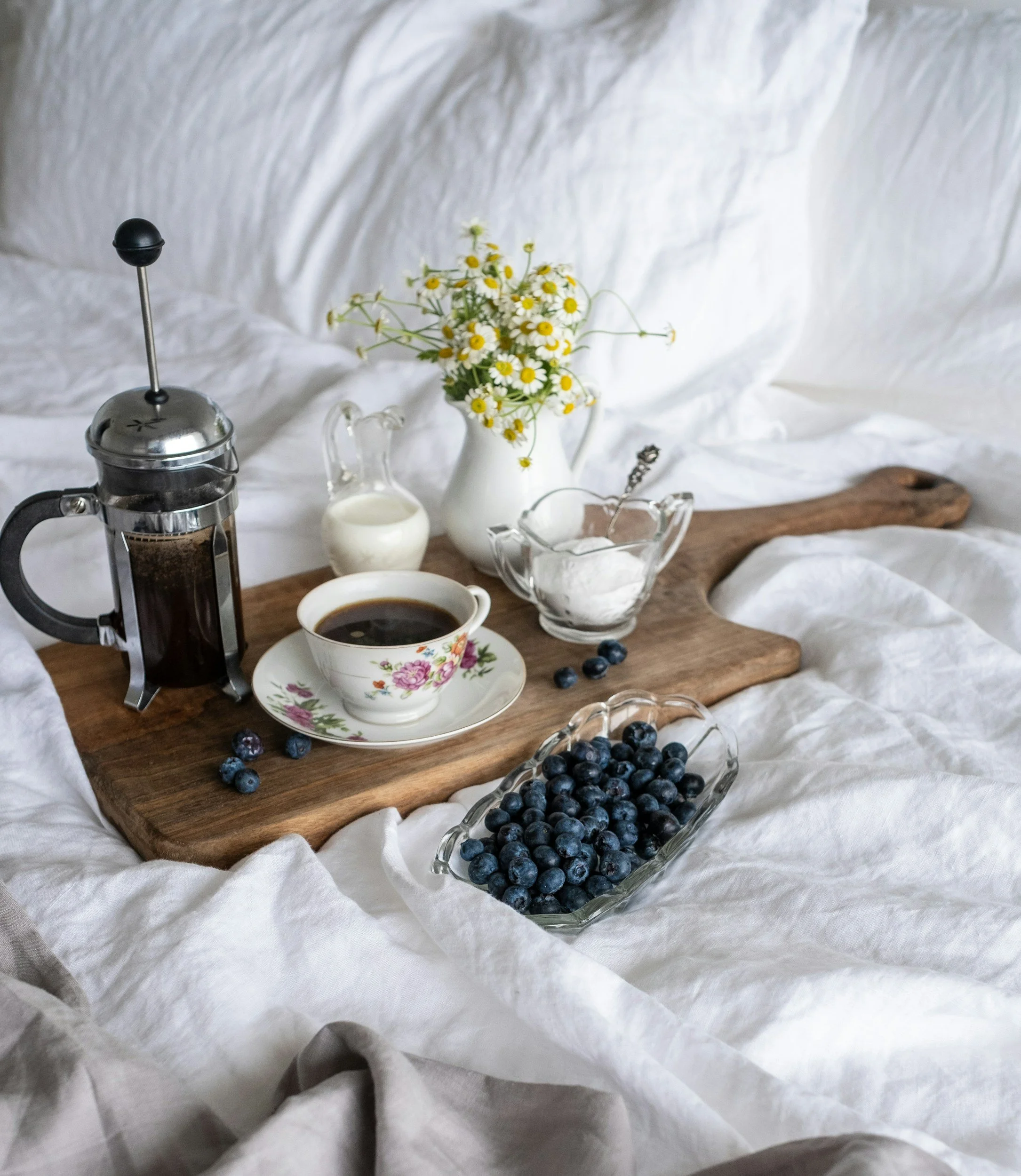 A breakfast scene with a French press, a cup of coffee, a small pitcher of cream, a bowl of blueberries, a jar of sugar, and a white vase with daisies, all placed on a wooden tray on white fabric.
