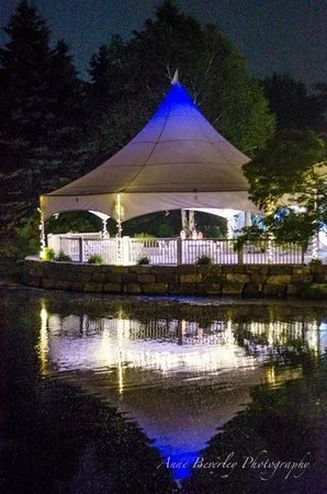 Nighttime view of a white gazebo with a pointed roof illuminated with blue lights, situated by a body of water with reflections, and surrounded by trees.