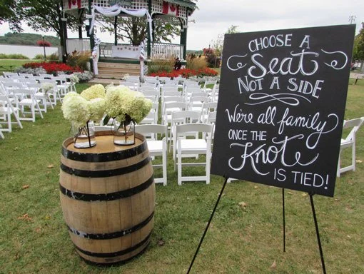 Chalkboard sign at outdoor wedding venue with seating instructions and a barrel topped with white hydrangea flowers.