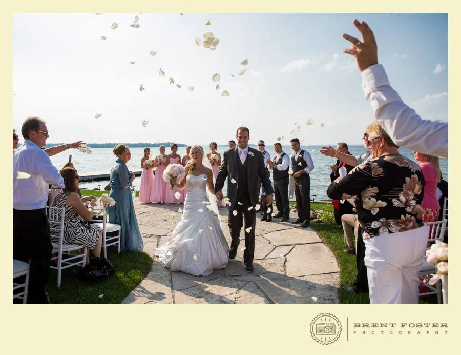 A bride and groom walk together down a stone path, surrounded by wedding guests throwing flower petals, with a lake and blue sky in the background.