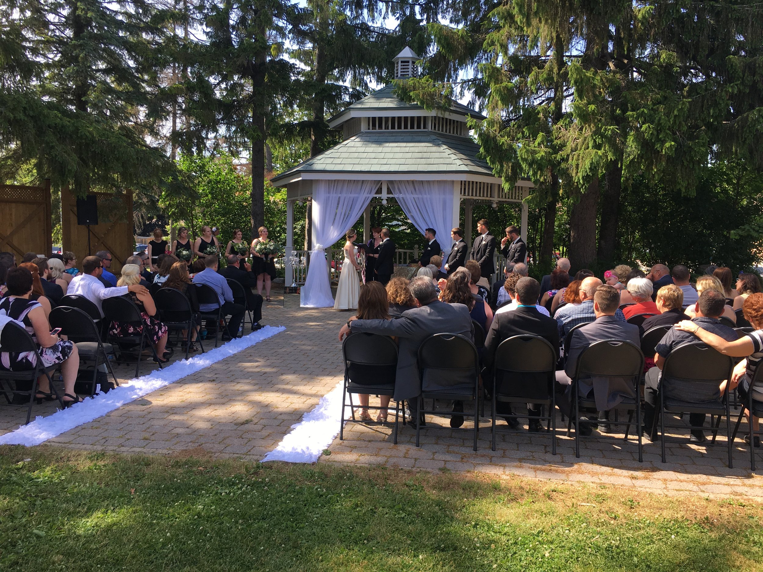 Outdoor wedding ceremony with a bride and groom standing under a gazebo, surrounded by bridesmaids and groomsmen, with guests seated on chairs facing the couple on a paved area with white decorative trim, in a garden setting with trees.