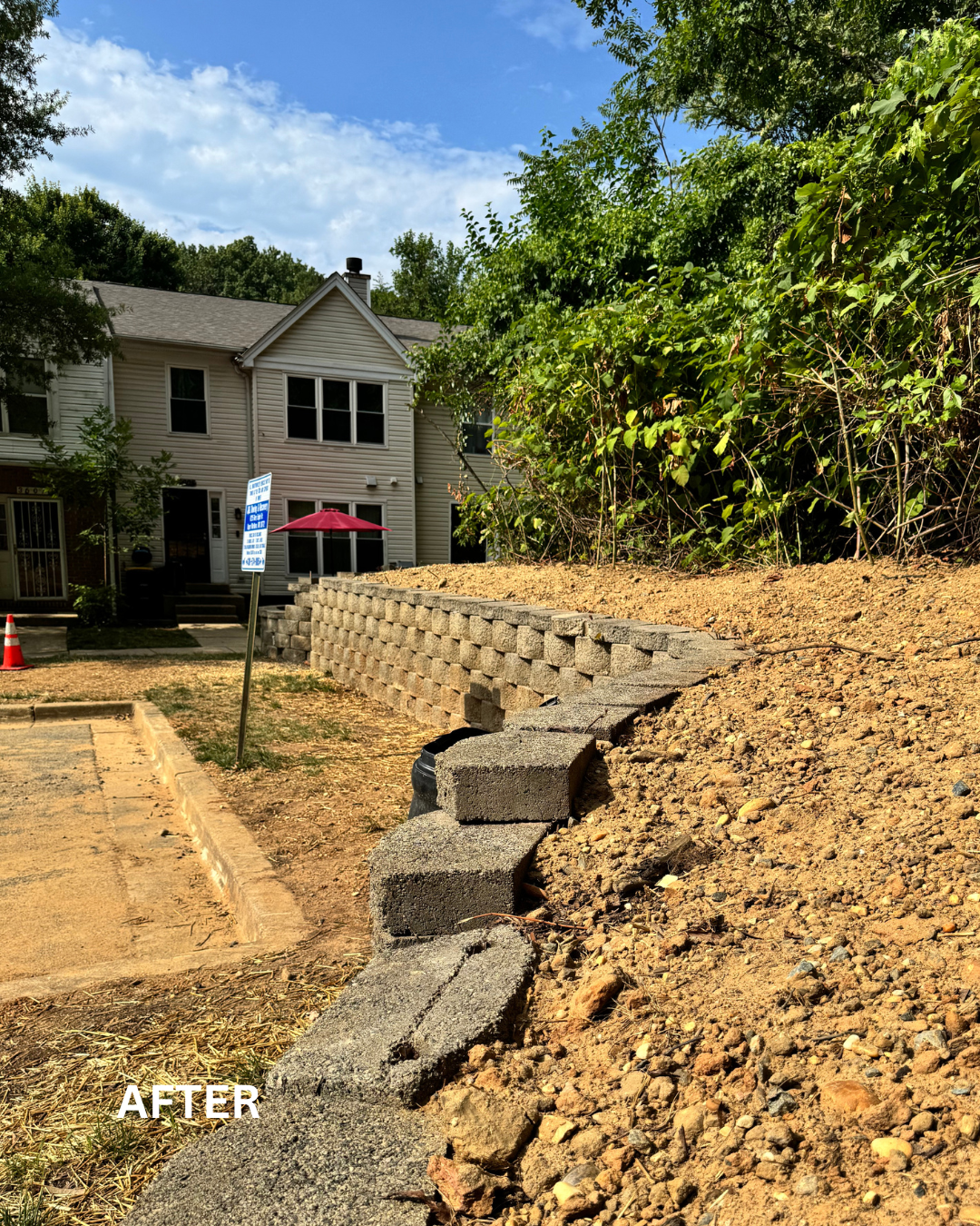 A new retaining wall of concrete blocks has been built along the edge of a slope behind a suburban house, with some loose concrete blocks and dirt in the foreground.