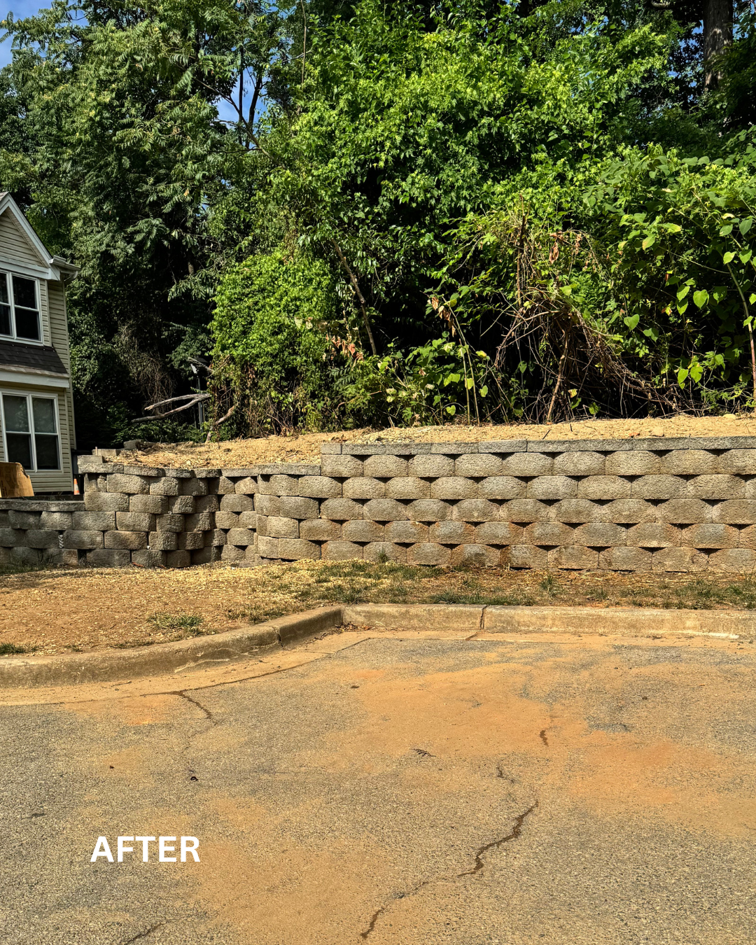 A retaining wall built with stacked concrete blocks supporting a hillside with green trees and bushes. A small section of a house is visible to the left. The ground in the foreground is cracked asphalt with a curb edge. The word 'AFTER' is written at