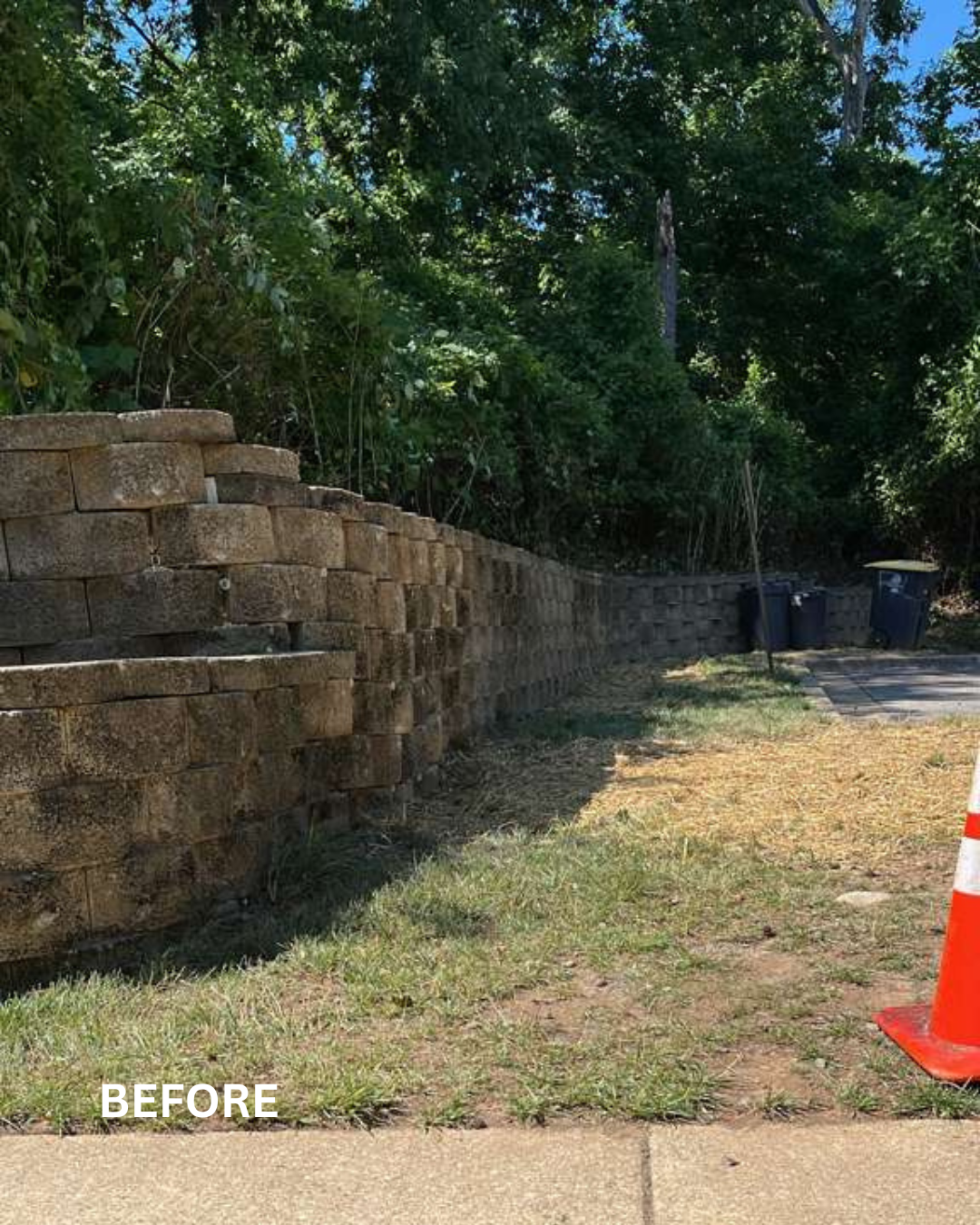 A curved brick retaining wall on the left side, with a grassy area and sidewalk in the foreground, two garbage bins, and a traffic cone on the right. Dense trees and greenery in the background.