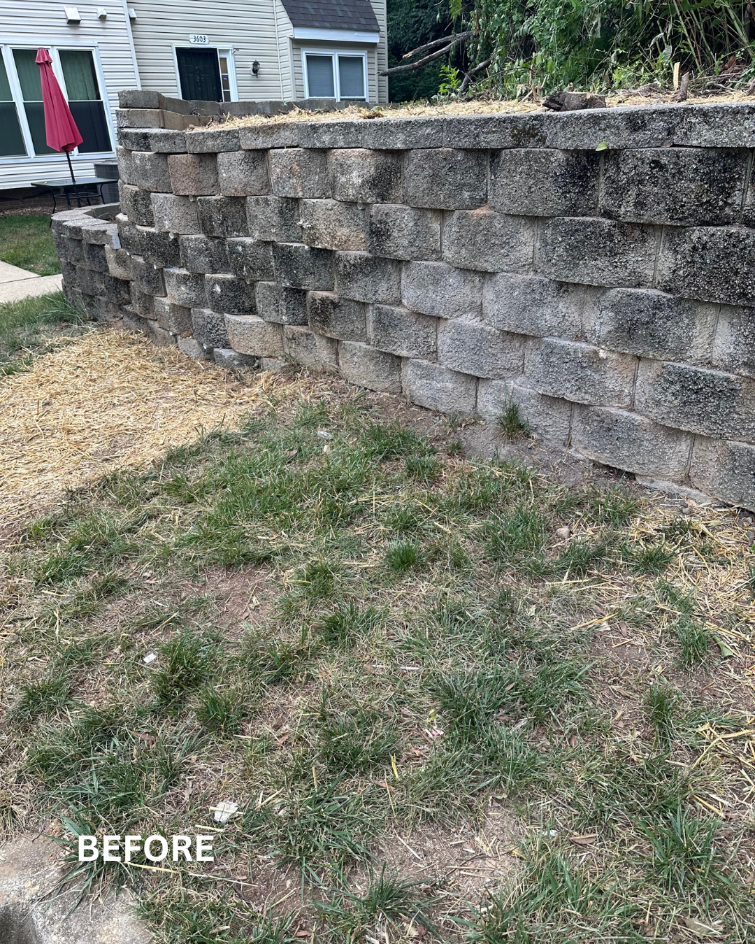 A retaining wall made of large gray stone blocks with some grass and straw in front, and a house with white siding and outdoor furniture behind.