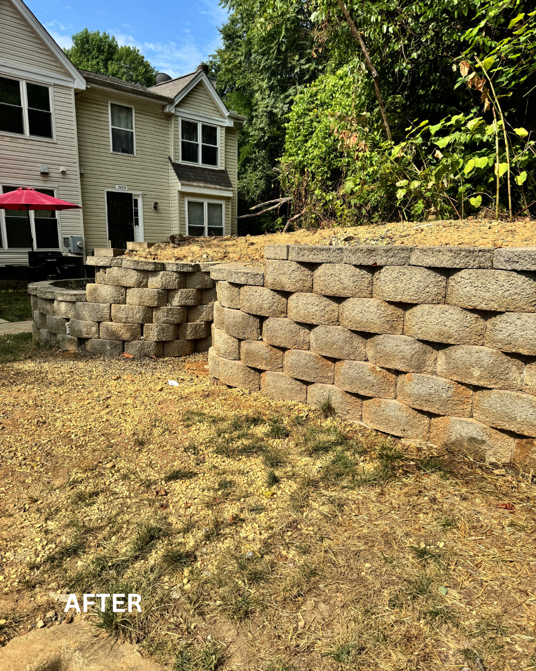 A newly constructed retaining wall made of beige stone blocks with a house in the background. The area in front of the wall has dirt and sparse grass, and there are trees and green foliage behind the wall.