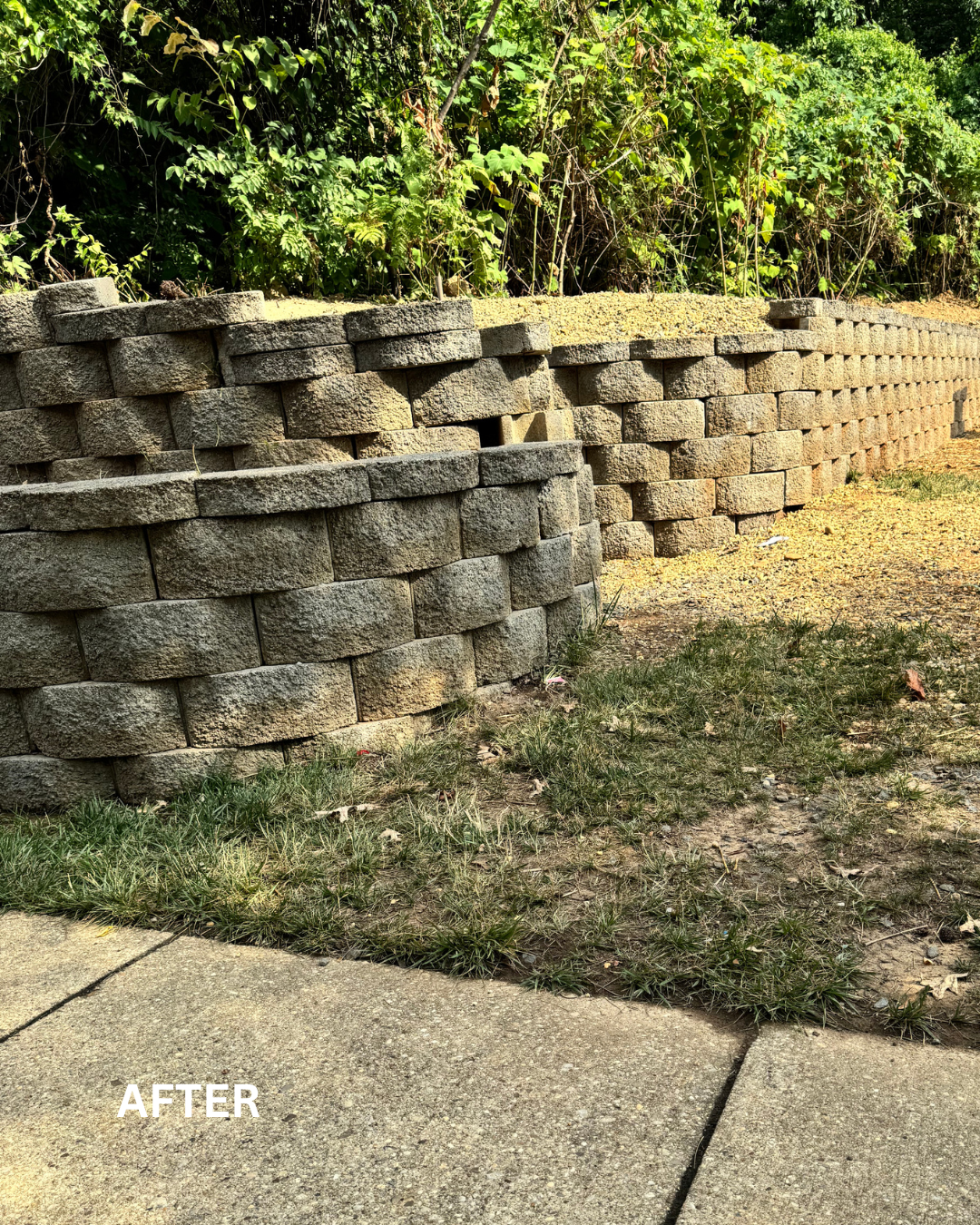 A newly constructed stone retaining wall with an uneven top, situated next to a sidewalk and overgrown grass and bushes.