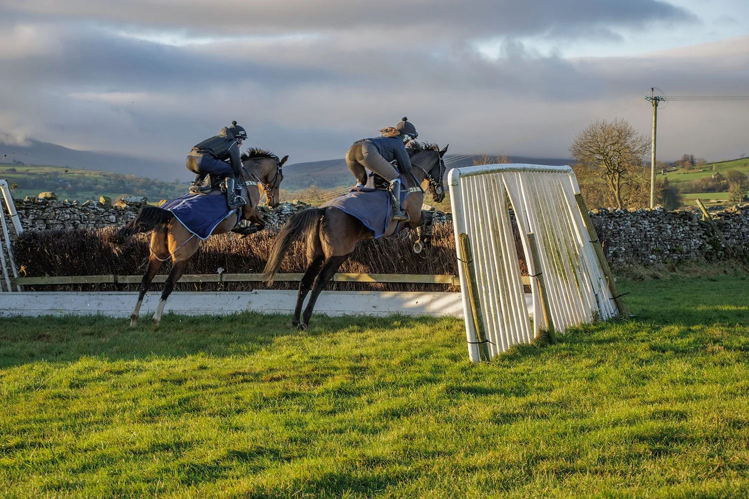 Two jockeys riding horses over a jump in a steeplechase race on a grassy field with hills and cloudy sky in the background.