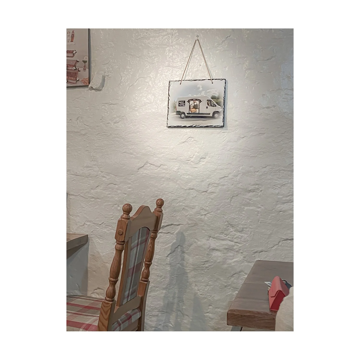 A wall with a textured, off-white surface, a framed picture hanging with a string, a wooden chair with a plaid cushioned seat, and a table with a small red napkin.