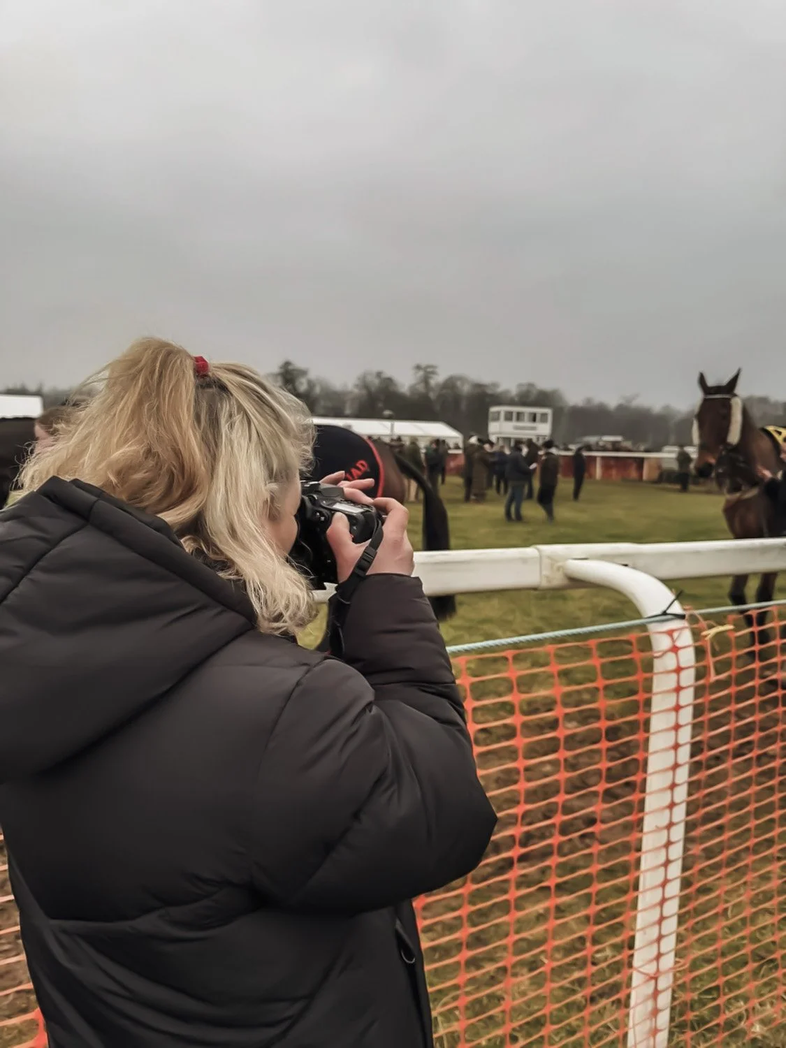 A woman with blonde hair in a black jacket is taking a photo with a camera at a horse race track, with horses and people in the background.