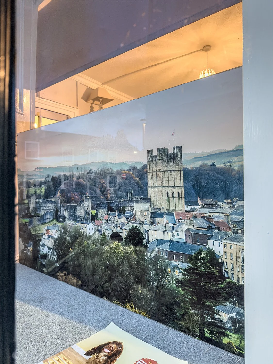 View of a town with a prominent castle tower on a hillside through a window, reflecting interior ceiling lights and a speaker.