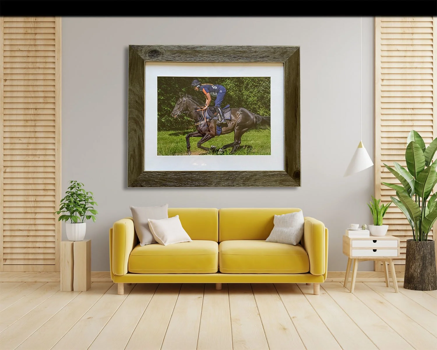 A living room with a yellow sofa, white pillows, a side table with a plant, another side table with a plant, and a large framed photo of a person riding a horse hanging on the wall.