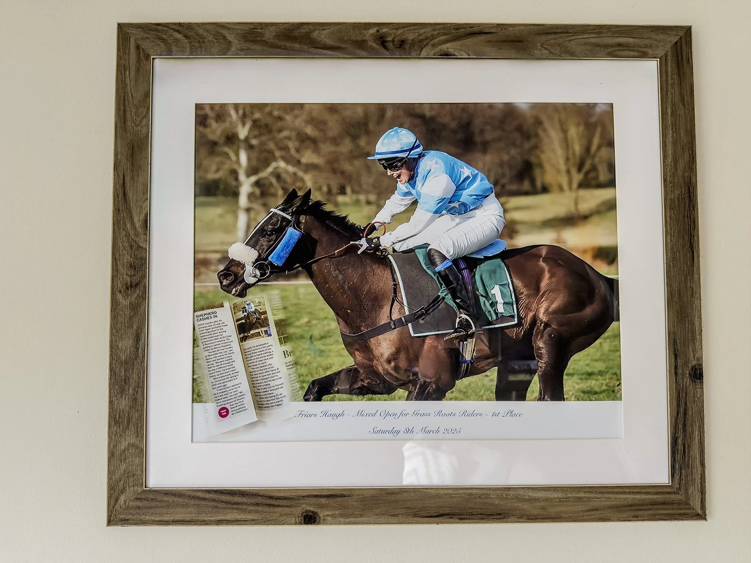 Photograph of a jockey riding a racehorse during a race, with a green field and trees in the background, framed and hung on a wall. A print by Tracey Jane's Photography and Prints