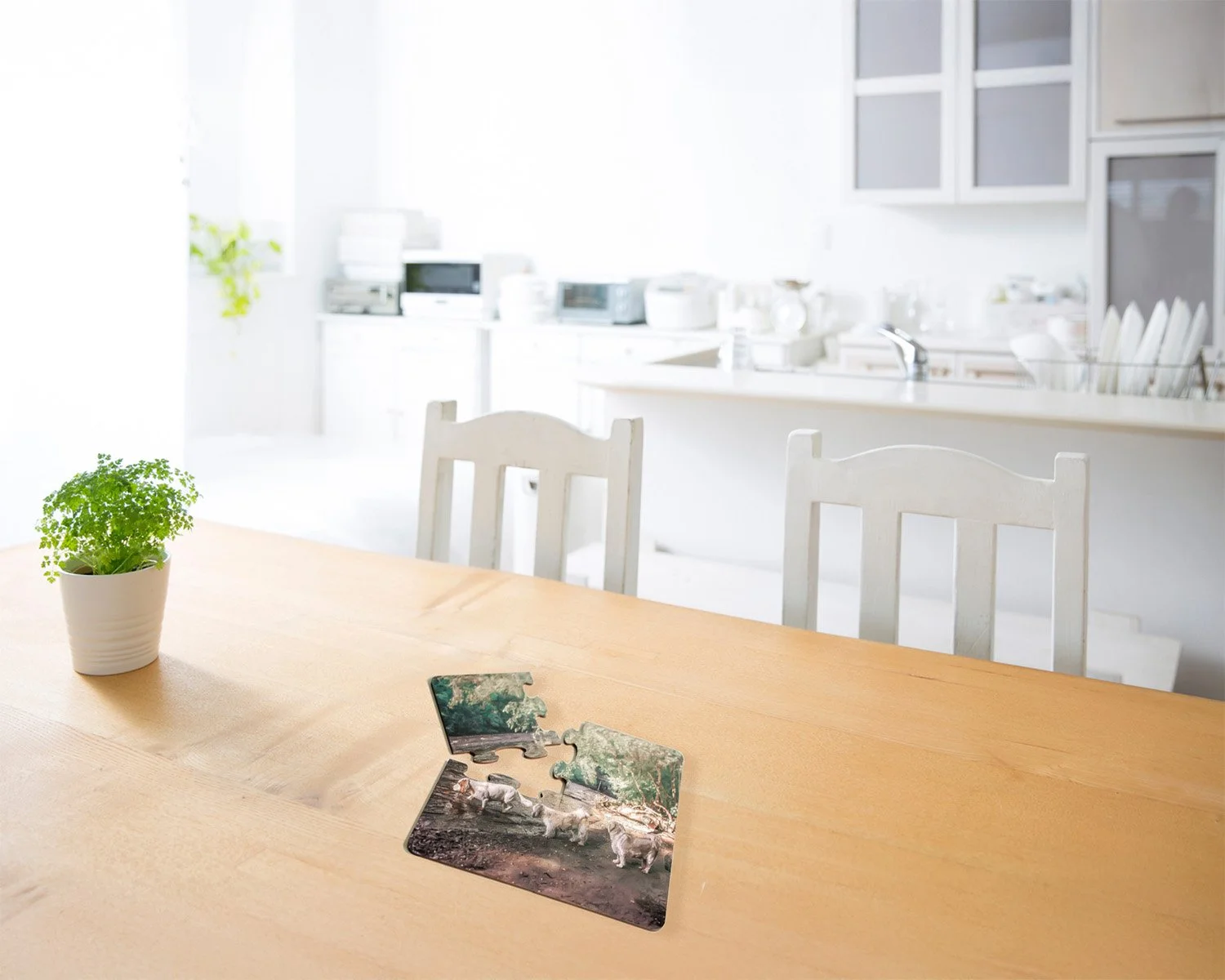 A light wooden dining table with a small potted green plant and two partially assembled jigsaw puzzles, set in a bright, white kitchen with white chairs and kitchen appliances in the background.