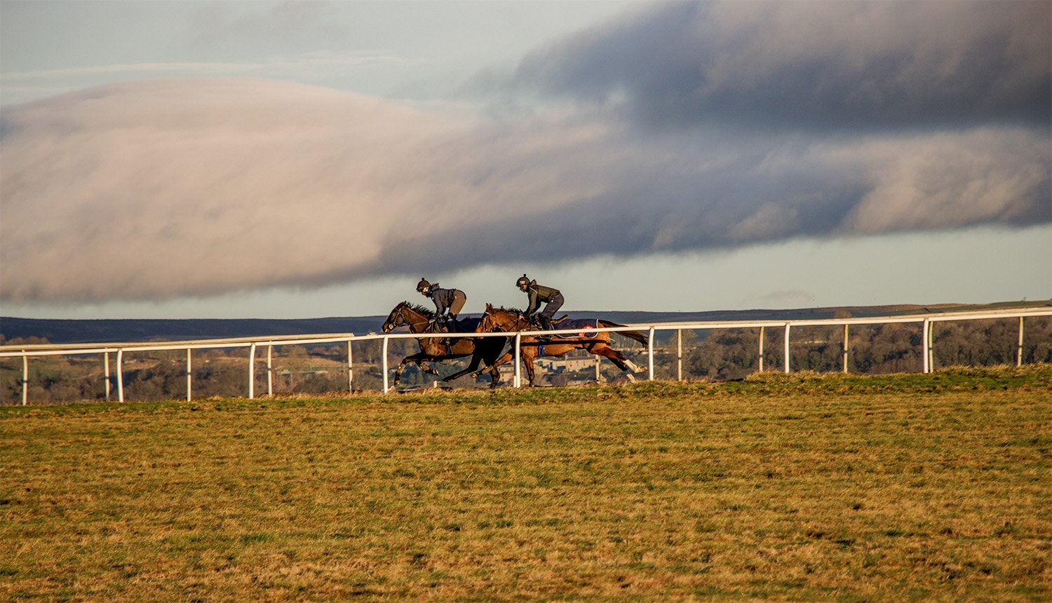 Two jockeys riding racehorses on the gallops with a cloudy sky in the background.