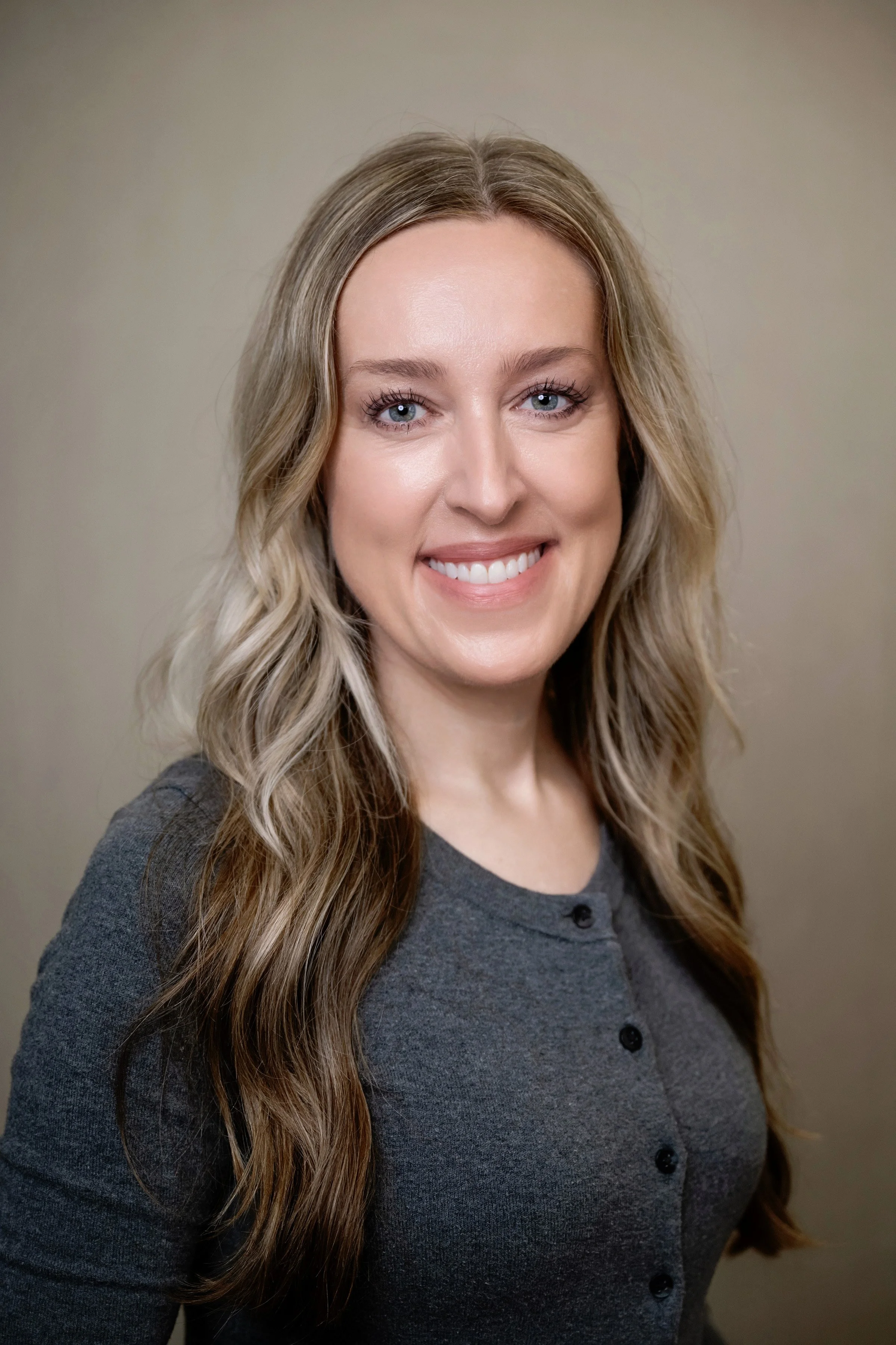 A woman with long blonde hair, blue eyes, wearing a light blue sweater, gold earrings, and a gold necklace, smiling against a dark blue background.