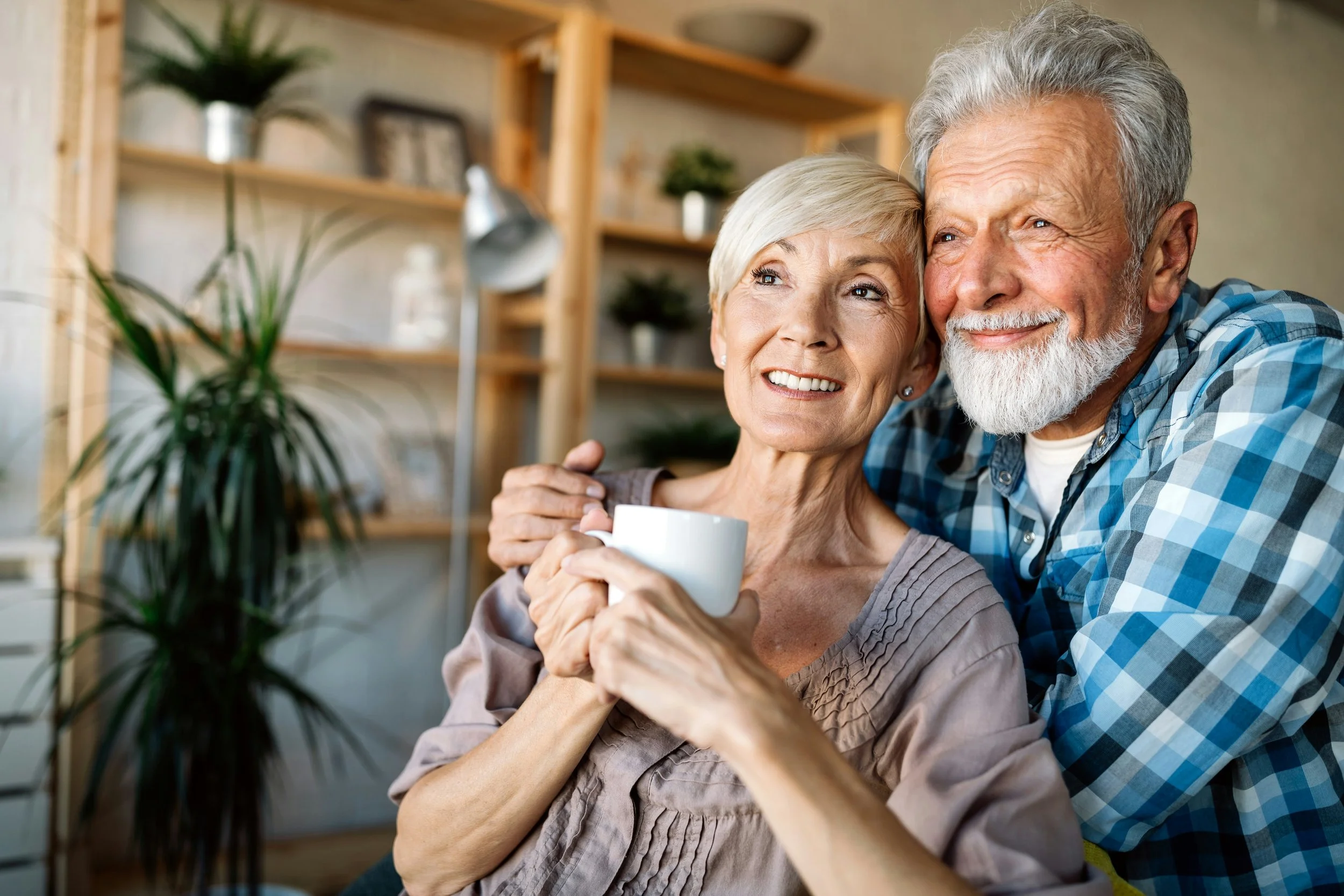 An elderly couple sitting close together, smiling, with the woman holding a white mug. The man has his arm around her.