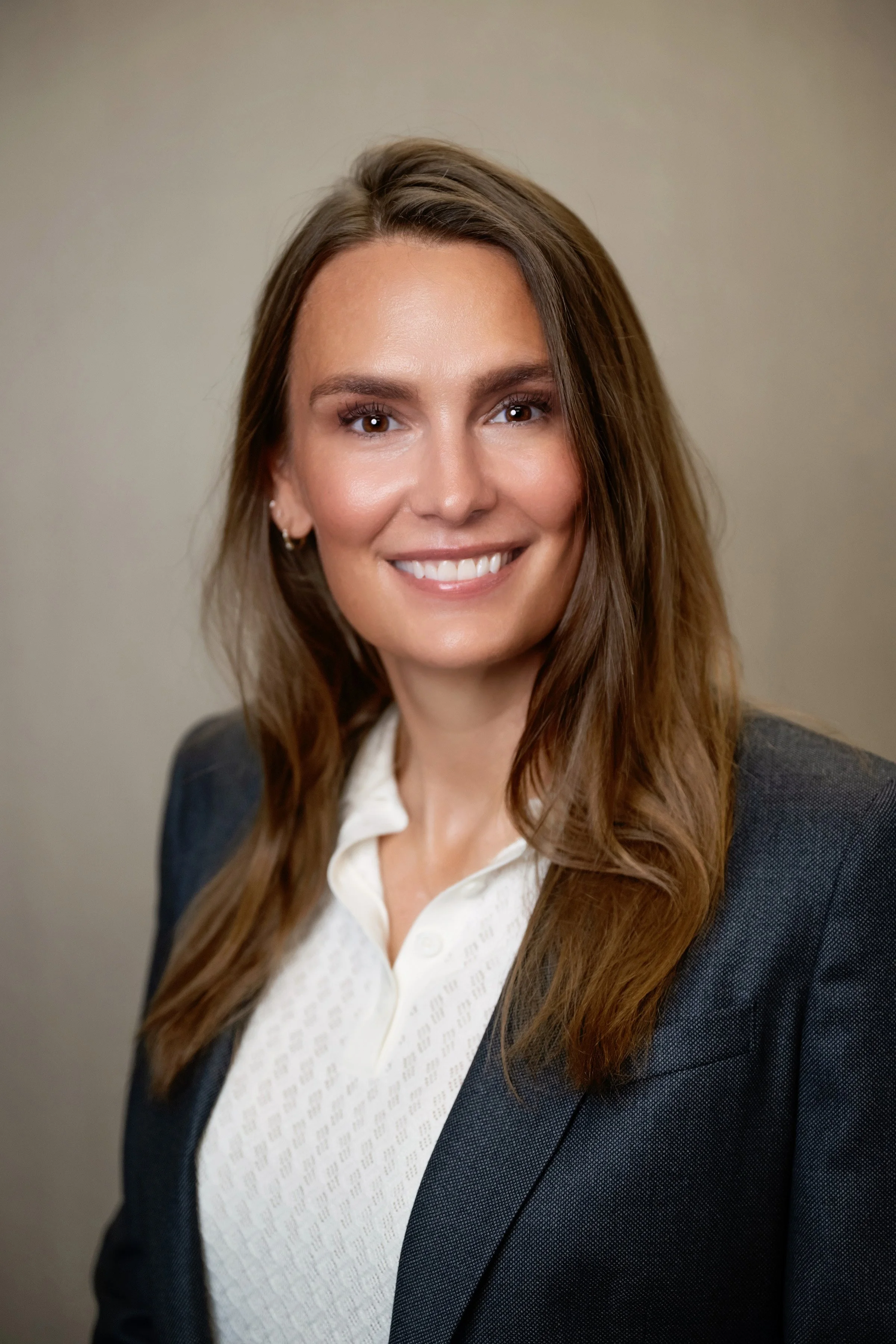 Close-up of a smiling woman in business attire, with long brown hair, wearing a white shirt and navy blazer, against a dark background.