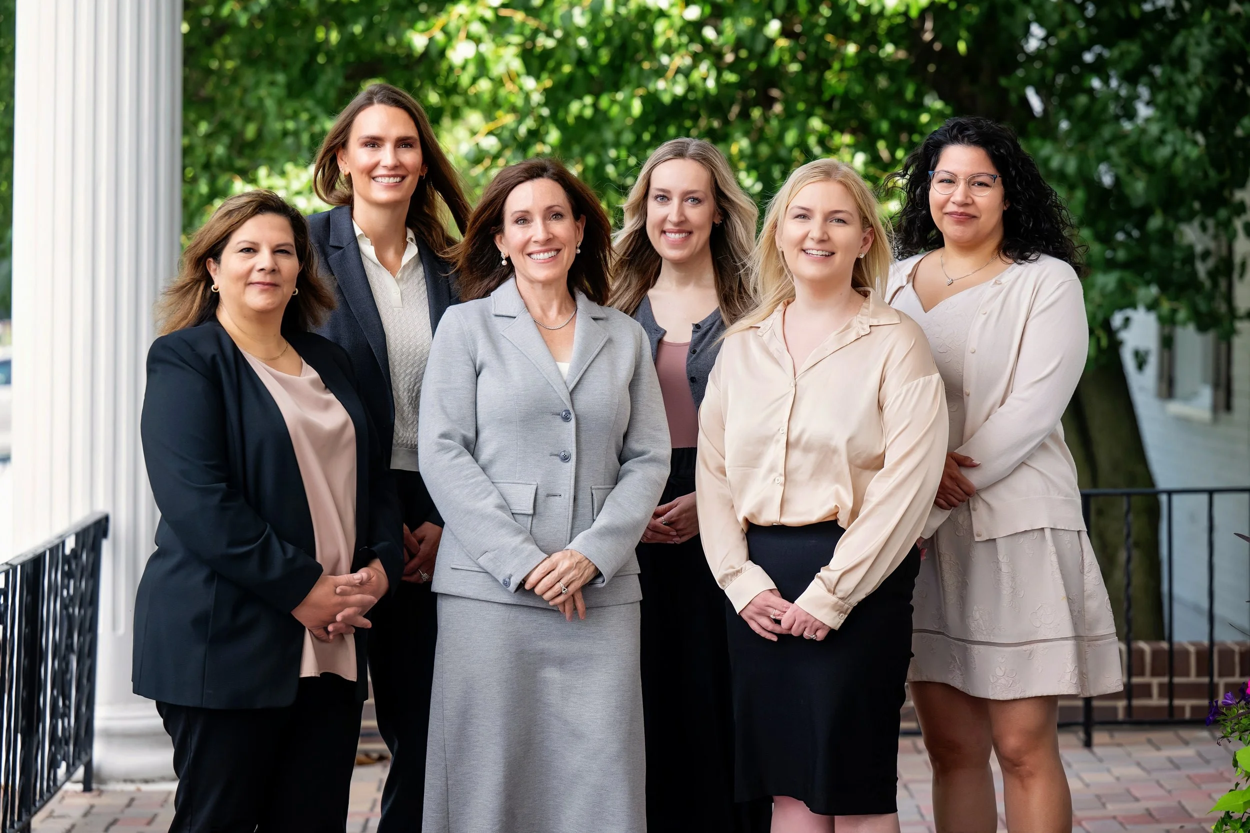 Group of seven women standing outdoors in front of green trees and a building, dressed in business casual attire, smiling for the photo.