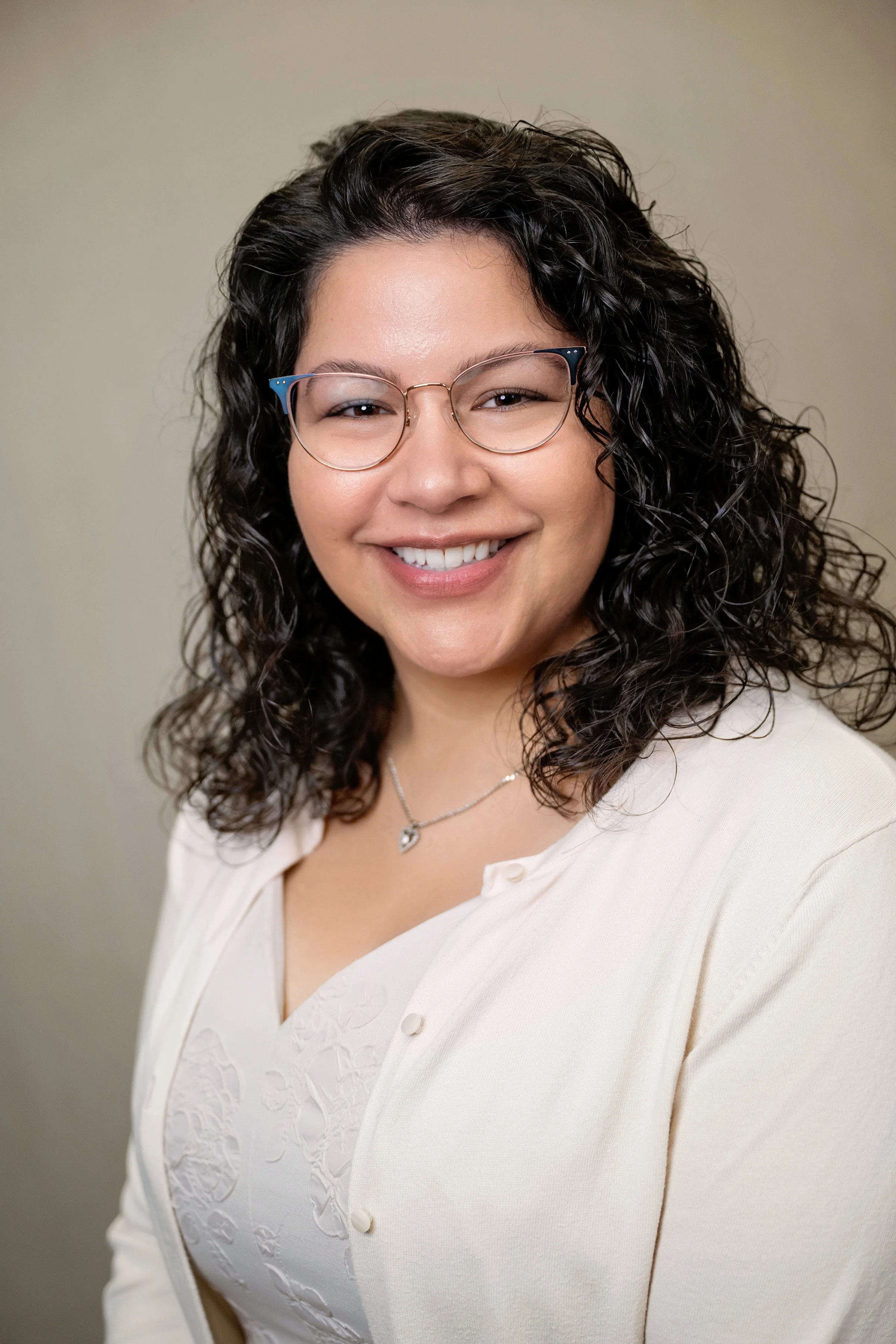 A woman with black, curly hair and glasses smiling against a dark blue background.