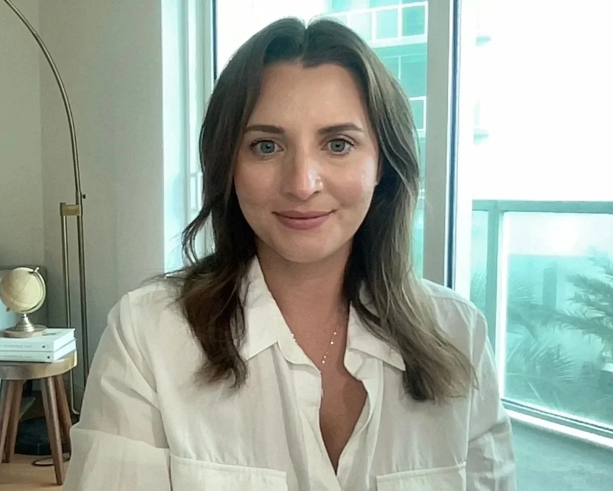 A woman with shoulder-length brown hair and blue eyes, wearing a white shirt, sitting indoors near large windows, with a background including a modern interior space and a small table with a globe and books.