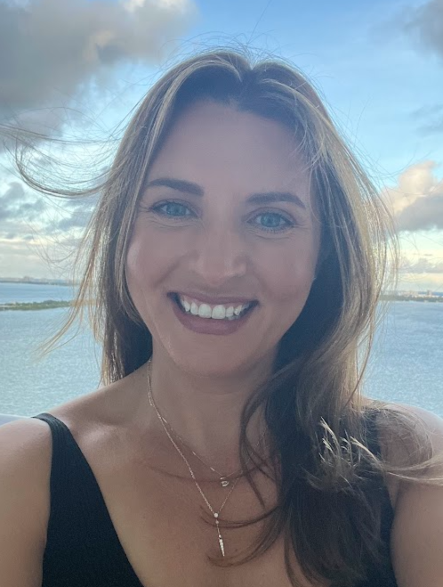 A woman with long brown hair and blue eyes smiling outdoors near a body of water against a cloudy sky.