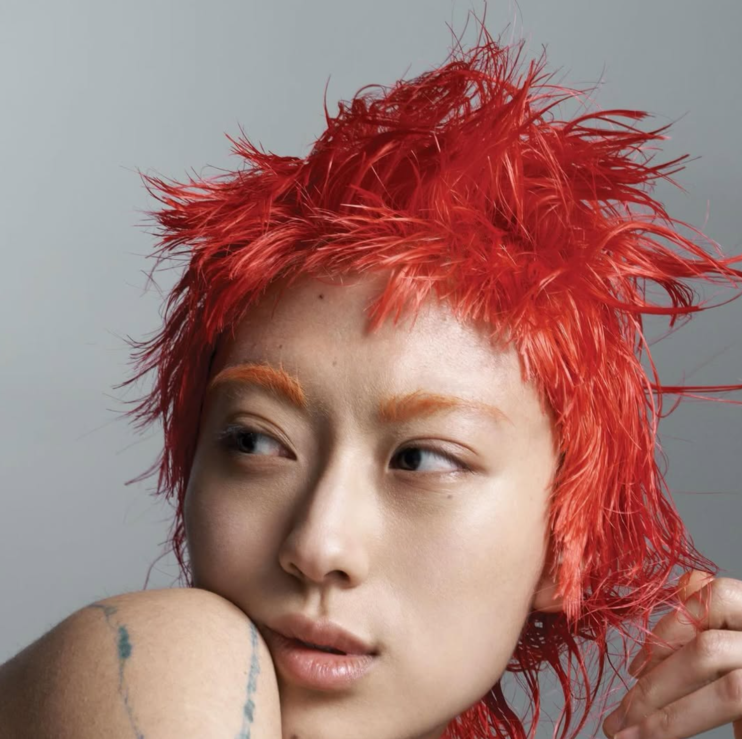 A close-up portrait of a person with wet, spiky, bright red hair, light skin, and light-colored eyebrows, looking to the side with a neutral expression, against a plain gray background.