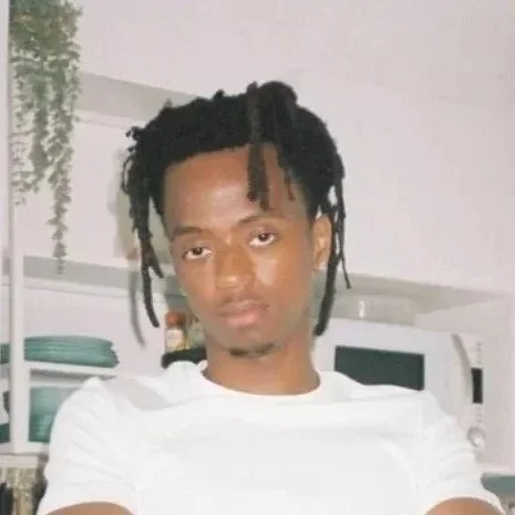 Young man with dreadlocks sitting on a chair against a brick wall, wearing a white t-shirt and light-colored pants, with a pink bag slung over his shoulder.