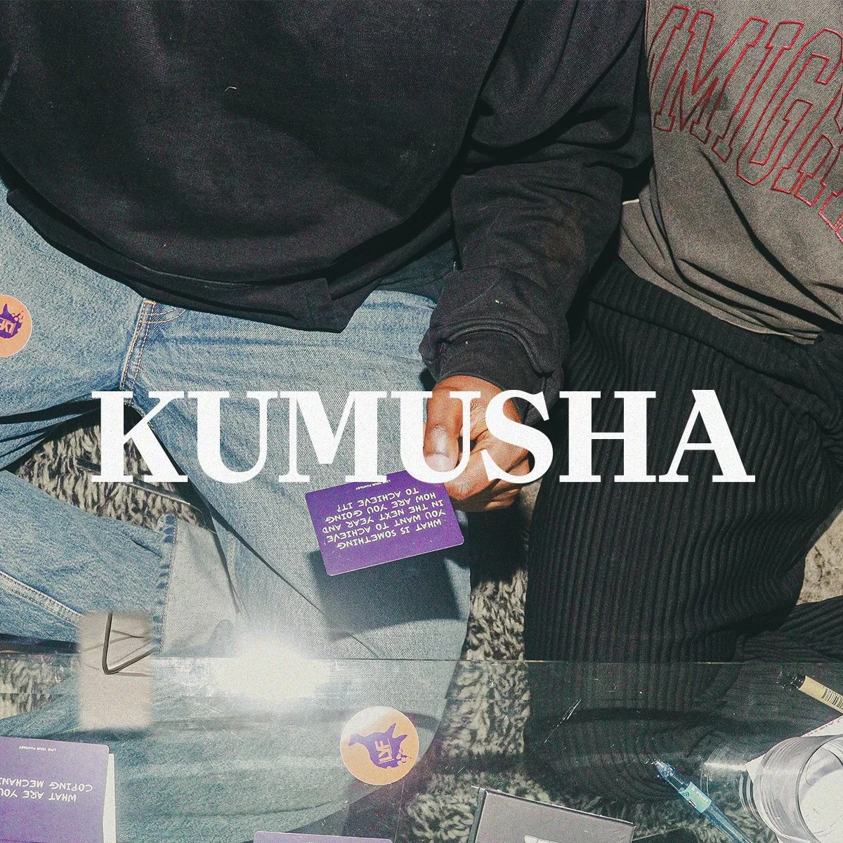 Close-up of two people sitting together, one holding a purple card with white text. The individuals are wearing dark and light clothing, jeans, and jackets, with a reflection showing on a glass surface. The word "KUMUSHA" is overlaid in large white letters on the image.