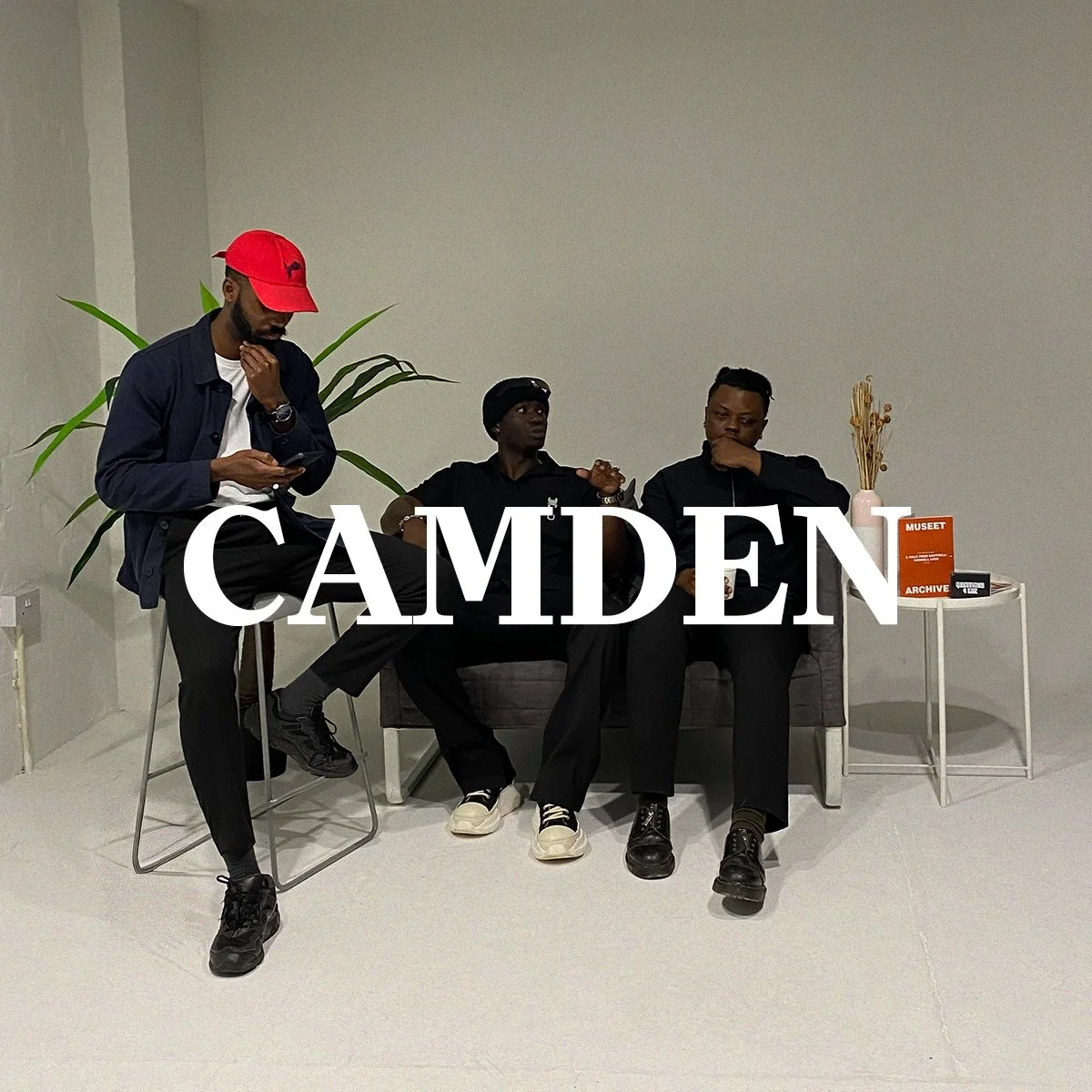 Three men sitting and standing in a minimalist room with a white wall. One man is standing on a stool, looking at his phone, while the other two are seated on a sofa, engaged in conversation. There is a large green plant behind them and a side table with a vase and a book titled 'Museet'.