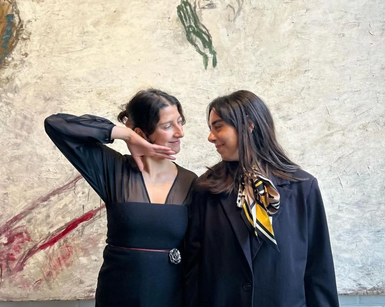 Two women face each other closely, one with short dark hair and the other with long dark hair, in front of an abstract textured wall.