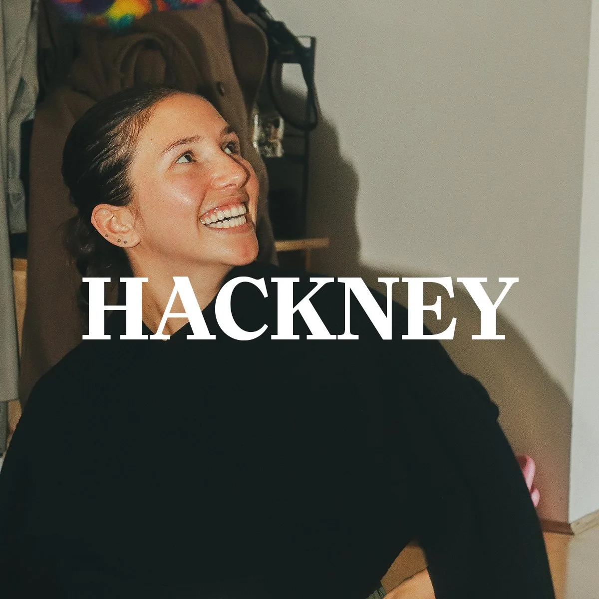 A woman with short dark hair smiling and looking upwards, sitting on the floor with her arms around her knees, in a room with a brown backpack and shelves in the background. The word "HACKNEY" is overlaid on the image.