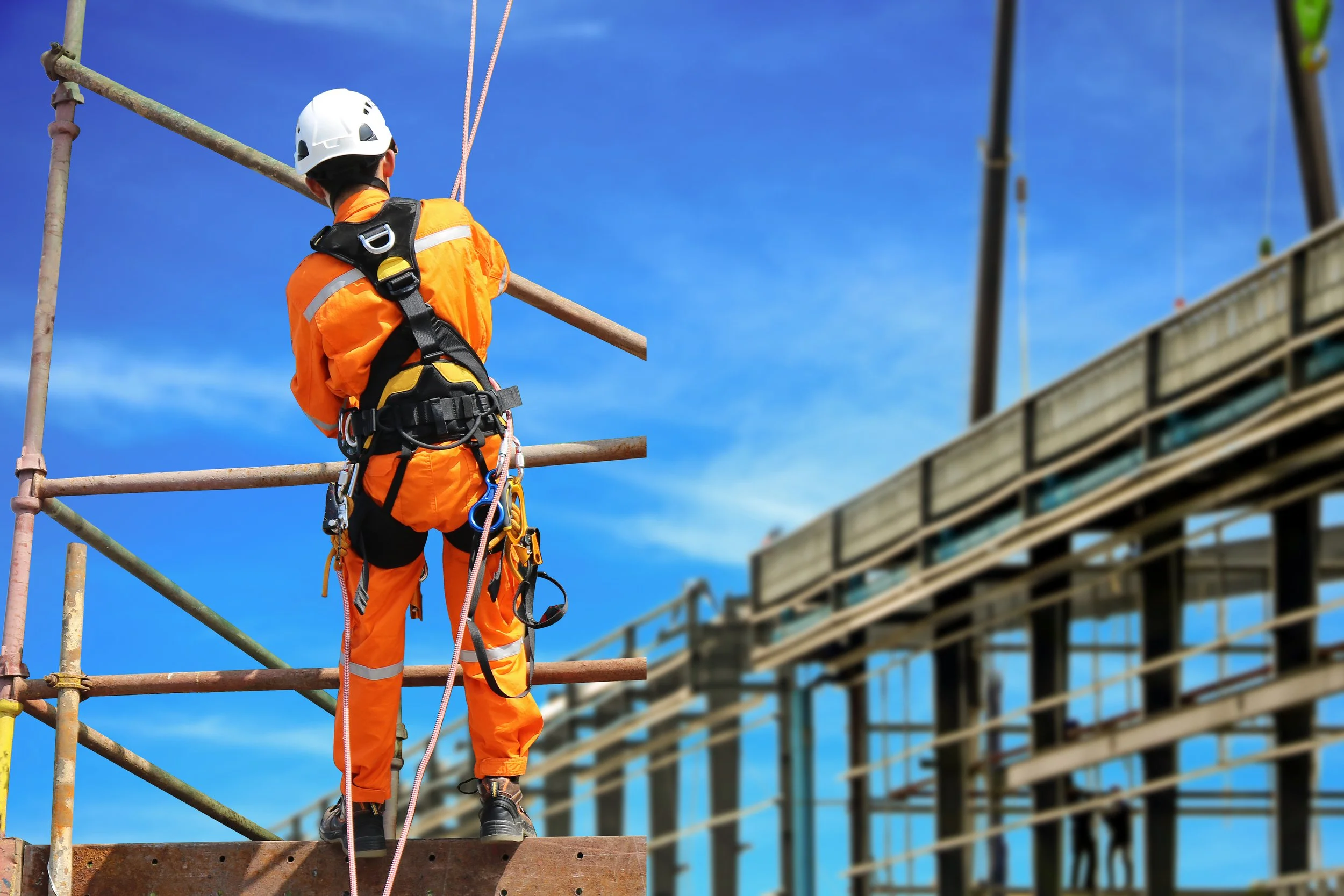 Construction worker in orange safety gear and helmet working on scaffolding at a construction site with a blue sky background.
