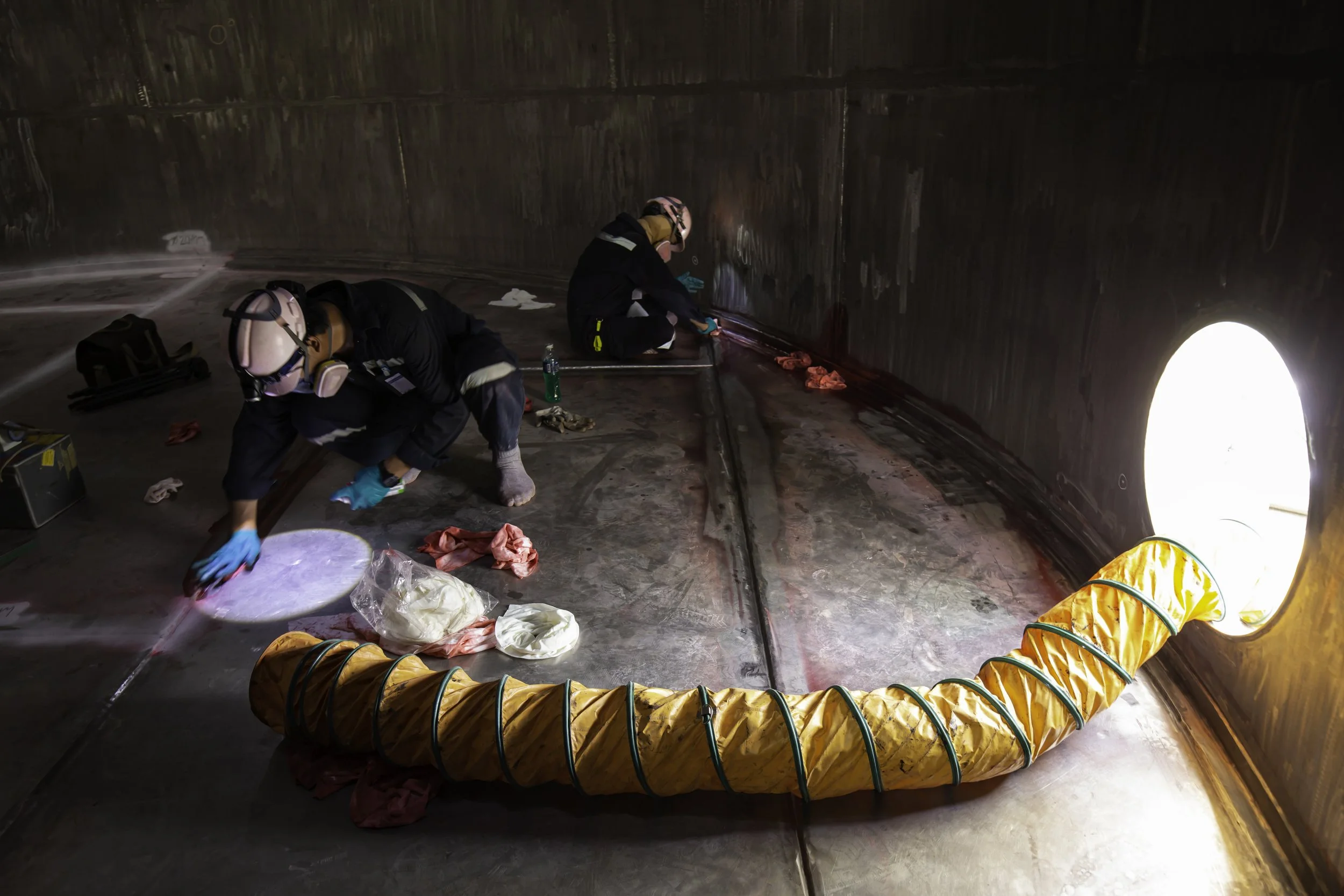 Two workers wearing protective gear working inside a large, circular industrial tank with a flexible orange hose connected to an opening. They are cleaning or inspecting the interior surfaces.